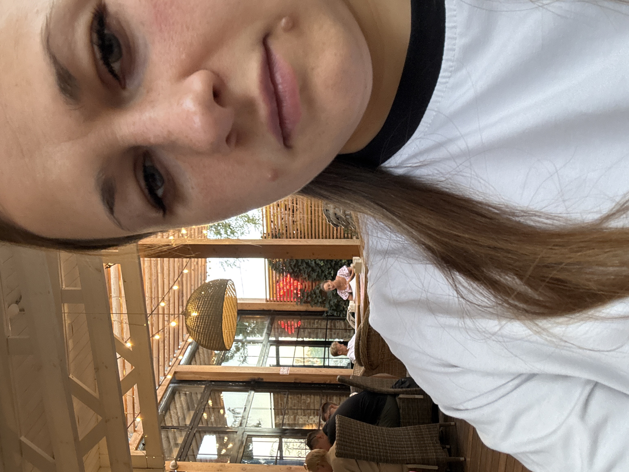 The image shows a close-up of a woman's face and shoulder in the foreground. She is wearing a white shirt with a black collar. In the background, an outdoor seating area of a restaurant is visible. There are tables and chairs, with other patrons seated. String lights are hung overhead, and a large woven lampshade is suspended from the ceiling. Large windows with wooden frames offer a view of greenery and a red illuminated sign. The woman's face is partially obscured by the camera angle, but her eyes, nose, and mouth are visible. There appear to be two small bumps on her chin. The scene suggests a casual dining environment. No specific location in Yevpatoriya, Ukraine is identifiable from the image.