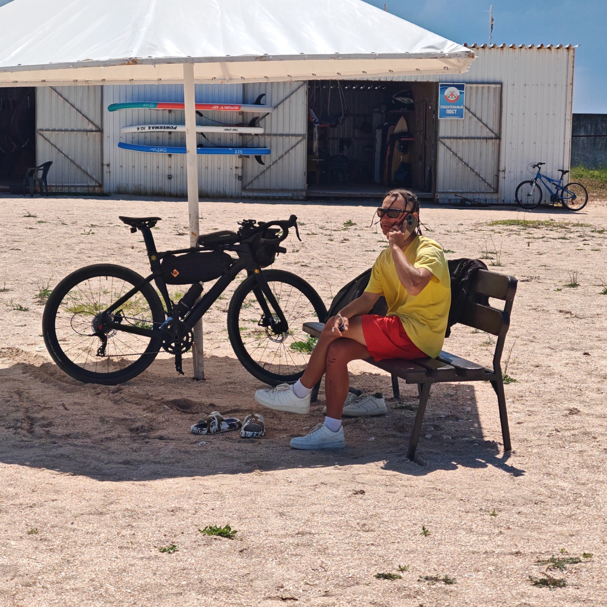 A man with dreadlocks, wearing sunglasses, a yellow t-shirt, and red shorts, is sitting on a wooden bench and talking on a mobile phone. He is wearing white sneakers and white socks. Next to him, on the sand, are a pair of patterned slides and another pair of white sneakers. A black bicycle with a black saddlebag is parked to his left. The bicycle has a black frame, black handlebars, and black wheels. A water bottle is attached to the bicycle frame. In the background, there is a white tent with a metal frame. Several paddleboards are mounted on the wall of a building behind the tent. The building is made of corrugated metal and has two large doors. A blue bicycle is parked to the right of the building. A sign with blue and red insignia and the text "СПАСАТЕЛЬНЫЙ ПОСТ" (Lifeguard Post) is visible on the building's wall. The ground is covered in sand and small shells. The scene appears to be outdoors in a coastal area. The location is identified as Мысовское сельское поселение, Ukraine.