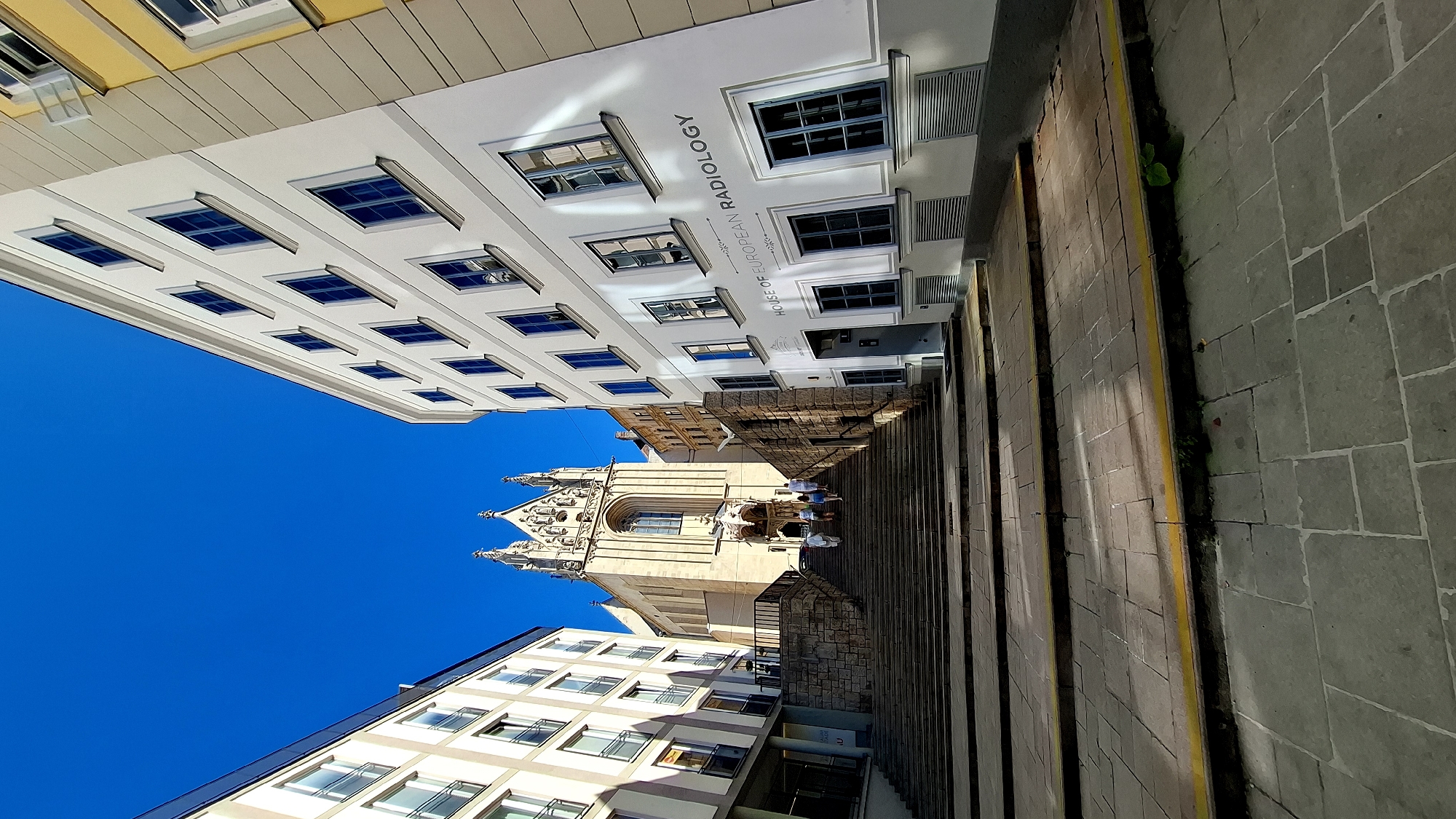 This image depicts a wide, stone staircase leading upwards towards a gothic-style building, possibly a church, with intricate architectural details and spires. The staircase is flanked by modern buildings on either side. The building on the right has a sign that reads "HOUSE OF EUROPEAN RADIOLOGY". Several people are walking up the stairs. The sky is a clear, bright blue, suggesting a sunny day. The scene is located in Vienna, Austria, specifically at the St. Stephen's Cathedral vicinity, near the House of European Radiology.