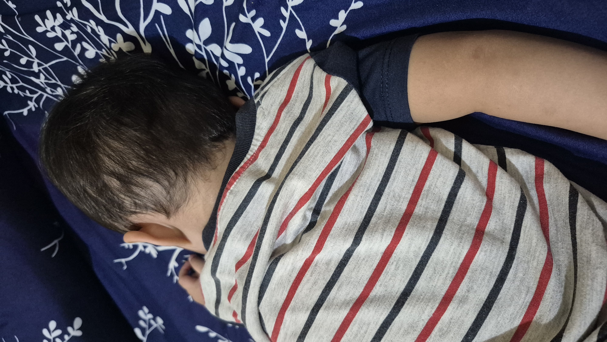 A young child is sleeping on a navy blue bed with a white floral pattern. The child is wearing a striped t-shirt with red, black, and white horizontal stripes. The child's back and head are visible. The environment appears to be an indoor bedroom. The image is captured from a top-down perspective. The scene is located in South Tangerang, Indonesia.