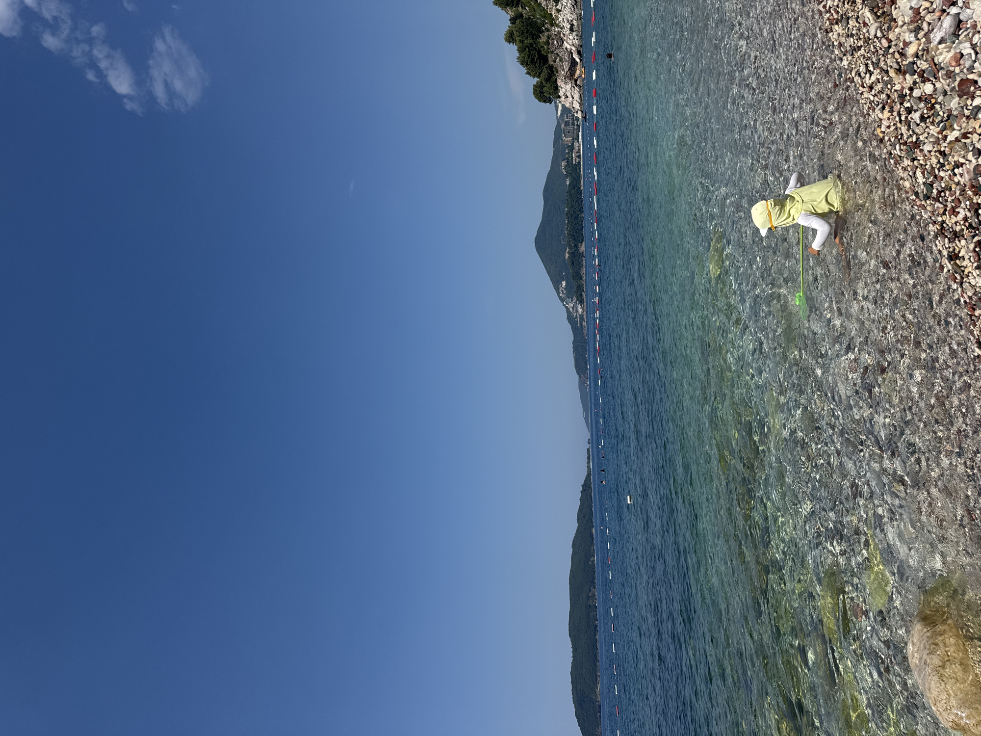 This media file shows a child playing on a pebble beach in Kamenovo, Montenegro. The child, wearing a yellow sun hat and a light-colored shirt, is bent over and holding a green net, seemingly exploring the shallow, clear water. The beach itself is composed of smooth, multi-colored pebbles.
The scene is set along a coastline with a gentle slope of trees and hills in the background. The sea is calm and a vibrant blue, with buoys marking a swimming area. The sky is a clear, deep blue, indicating sunny weather. The sun appears to be high in the sky, suggesting it is midday. The overall impression is one of a peaceful summer day by the sea, with a focus on the child's engagement with nature. There is no visible text in the media file.