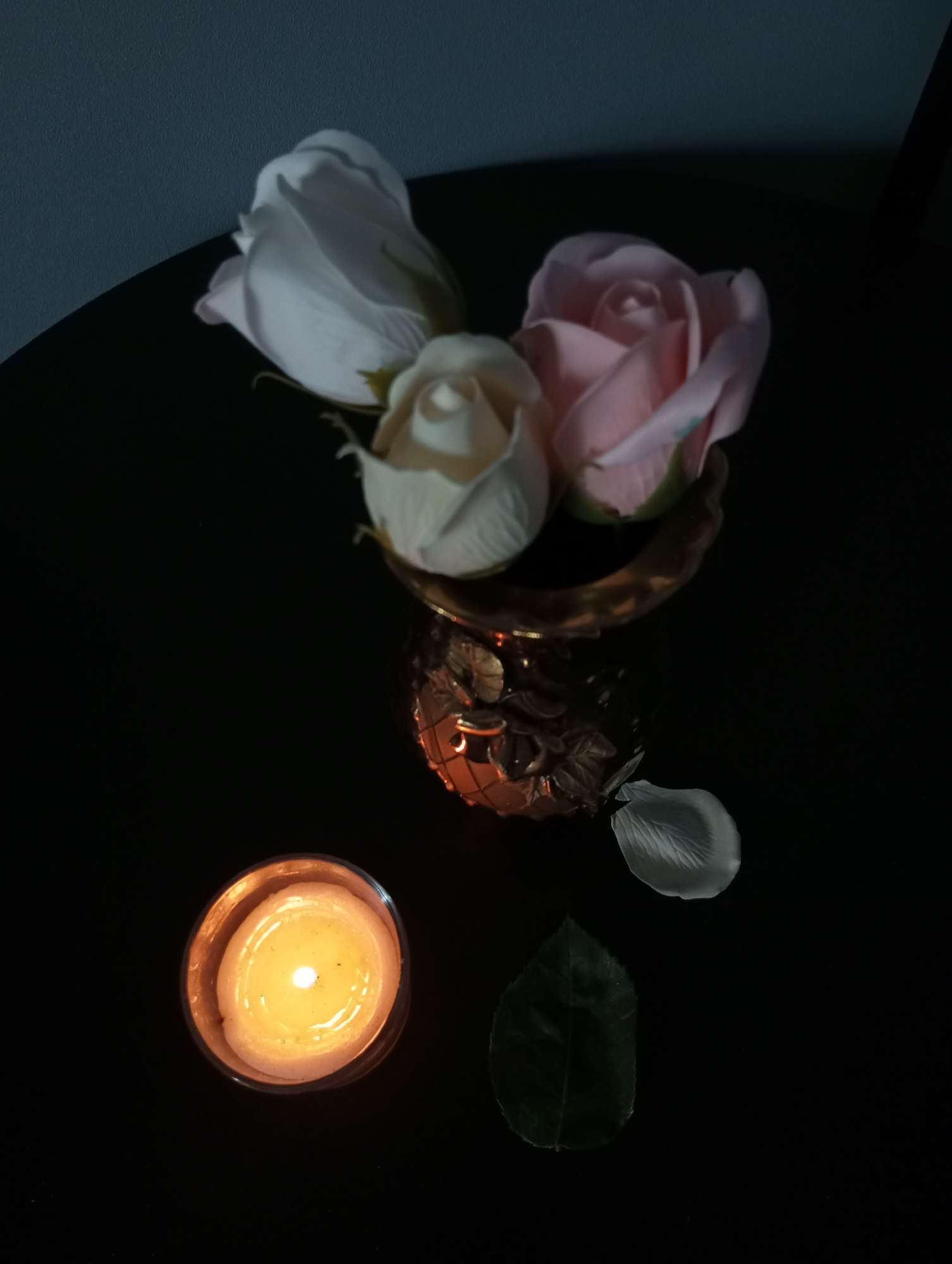 The image captures a dimly lit still life composed of artificial flowers and a burning candle on a dark, reflective surface. Three rosebuds, likely artificial, are arranged in a small, ornate metallic vase or holder in the upper center of the frame. From left to right, these include a white rose, a pale cream-colored rose, and a light pink rose, all mostly closed.
In the lower left quadrant, a single candle burns inside a glass holder, emitting a warm, golden light that illuminates the surrounding objects and casts a glow on the dark surface. To the right of the candle, a single, translucent white rose petal lies on the dark surface, and below it, a dark green leaf is visible. The background is predominantly dark, with a subtle hint of a dark blue or grey wall in the upper left corner.
The scene conveys a quiet and serene mood, suggesting an evening or nighttime setting due to the low light and the candle serving as the primary light source. There are no people visible, nor is there any visible text or ongoing activity, emphasizing the tranquil and contemplative nature of the arrangement.