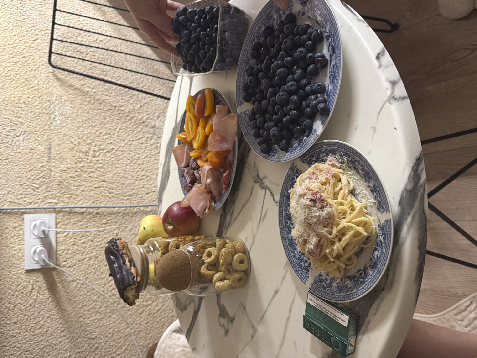 A close-up, eye-level shot captures a marble-top table set with a variety of food items and a power outlet visible in the background. On the left, a dark chocolate eclair sits atop a glass jar filled with ring-shaped crackers. Next to the jar are two apples, one red and one yellow. A plate of charcuterie, featuring sliced ham, salami, and cherry tomatoes, is situated to the right of the apples. Further to the right, two plates are filled with blueberries, one in a clear plastic container and the other on a patterned ceramic plate. In the foreground, a plate of spaghetti carbonara, generously topped with grated cheese, is positioned next to a green cigarette pack. The table's surface has a distinctive white and gray veined marble pattern. A partial view of a person's hand is visible reaching for the blueberries, and another person's leg is partially visible on the left. The background features a textured, light-colored wall with a white double electrical outlet and two plugs inserted. A portion of a black metal chair is visible on the right side of the frame. The overall scene suggests a casual meal or snack, likely indoors, with no specific indication of the city of Tbilisi, Georgia.