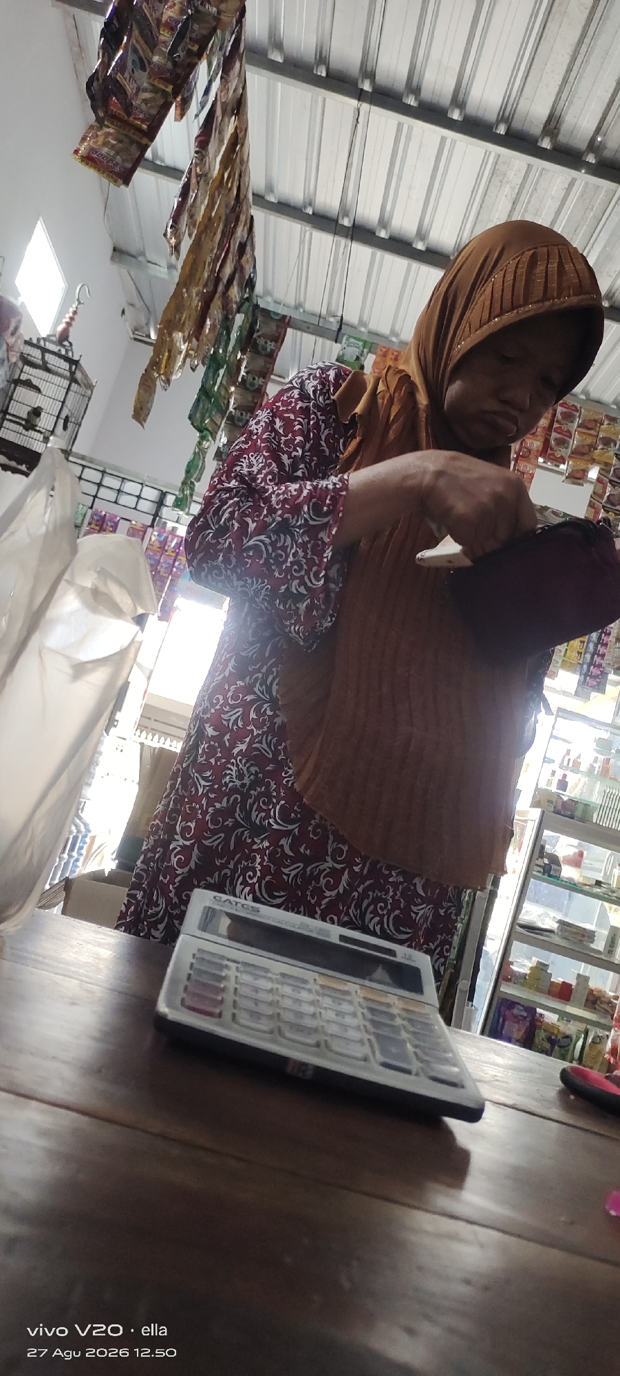 The image shows a woman in a hijab and a maroon and white patterned dress. She is standing behind a wooden counter in what appears to be a small shop. In front of her on the counter is a calculator. The woman is holding a brown, textured object in her hands, possibly a wallet or a small bag, and her attention is directed downwards. Behind her, shelves are visible with various items, suggesting a retail environment. Hanging from the ceiling are rows of packaged goods, possibly snacks or candy. To the left of the woman, a birdcage with a bird inside is visible, and a large white plastic bag is also present. The overall lighting suggests an indoor setting with some natural light coming from the right. The timestamp "27 Agu 2026 12.50" is present in the bottom left corner, indicating a future date. This scene likely depicts a transaction or an inventory check in a local shop in Simo, Indonesia.