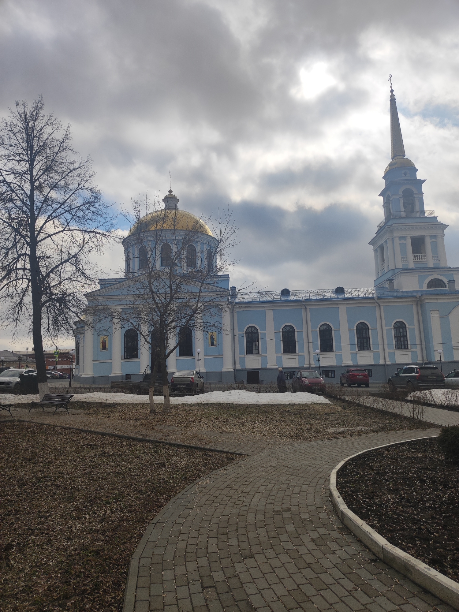 The image shows a light blue and white Orthodox church with a golden dome and spire in Votkinsk, Russia. Cars are parked near the church. A paved walkway leads through a park-like area with bare trees, dead grass, and patches of snow. The sky is overcast.