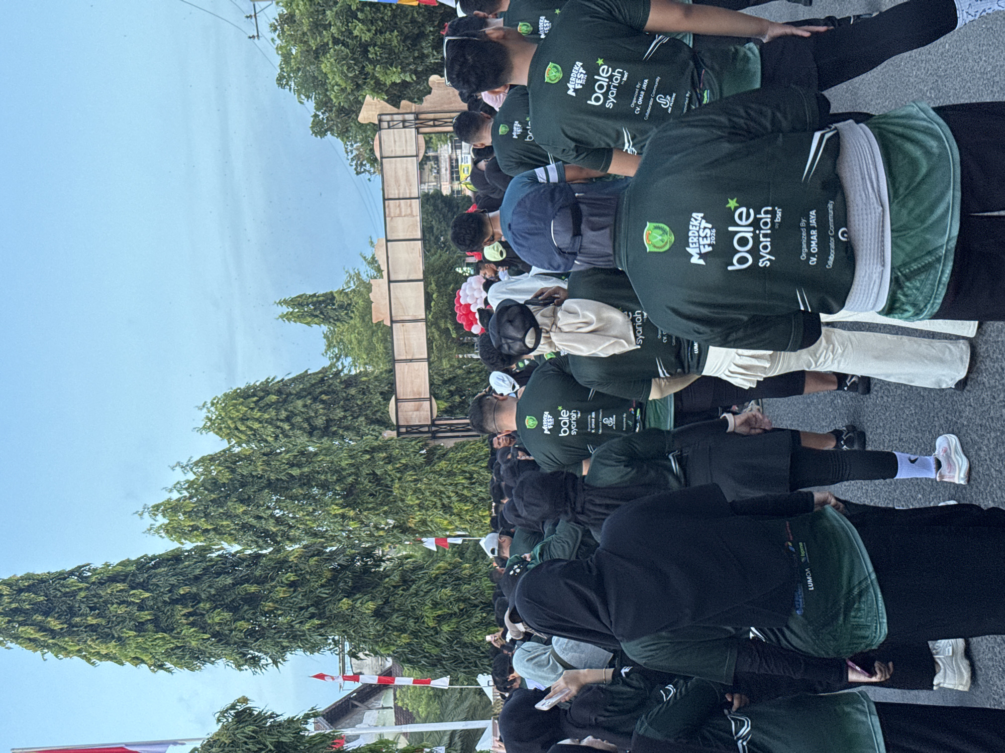 The image shows a crowd of people gathered outdoors, likely for an event. Many individuals are wearing dark green t-shirts with white lettering that reads "MERDEKA FEST 2026 bale syariah". Some t-shirts also have "Organized by: CV. OMAR JAYA" and "Collaborator Community" printed on them. The crowd appears to be moving forward, possibly towards a large wooden archway in the background. Flags, including Indonesian flags, are visible in the upper left portion of the image, suggesting a national or local celebration. Tall, lush green trees are prominent throughout the scene. The ground is paved, indicating an urban or semi-urban setting. The sky is a light blue with some wispy clouds. The overall atmosphere seems to be one of community gathering and participation. The presence of "Lhokseumawe, Indonesia" in the context of the image points to this city as the location of the event.