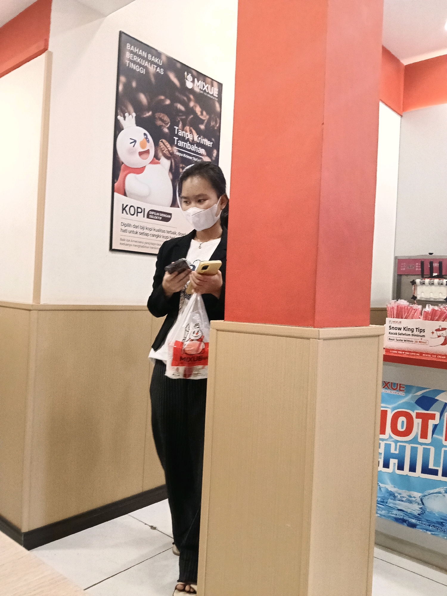 A woman wearing a white face mask and black jacket over a white t-shirt is standing indoors, holding two smartphones and a white plastic bag with red graphics. She appears to be looking at her phones. The environment suggests a retail setting with a large poster featuring coffee beans and a cartoon character on the wall behind her. To her right, there is a large red pillar and a display shelf with pink straws and a sign that reads "Snow King Tips". Further to the right, a sign with the word "XUE" and "HOT CHILLI" is visible. The flooring appears to be white tile. The woman's attire and the presence of retail displays suggest she is likely in a shop or cafe in Belimbing, Indonesia.