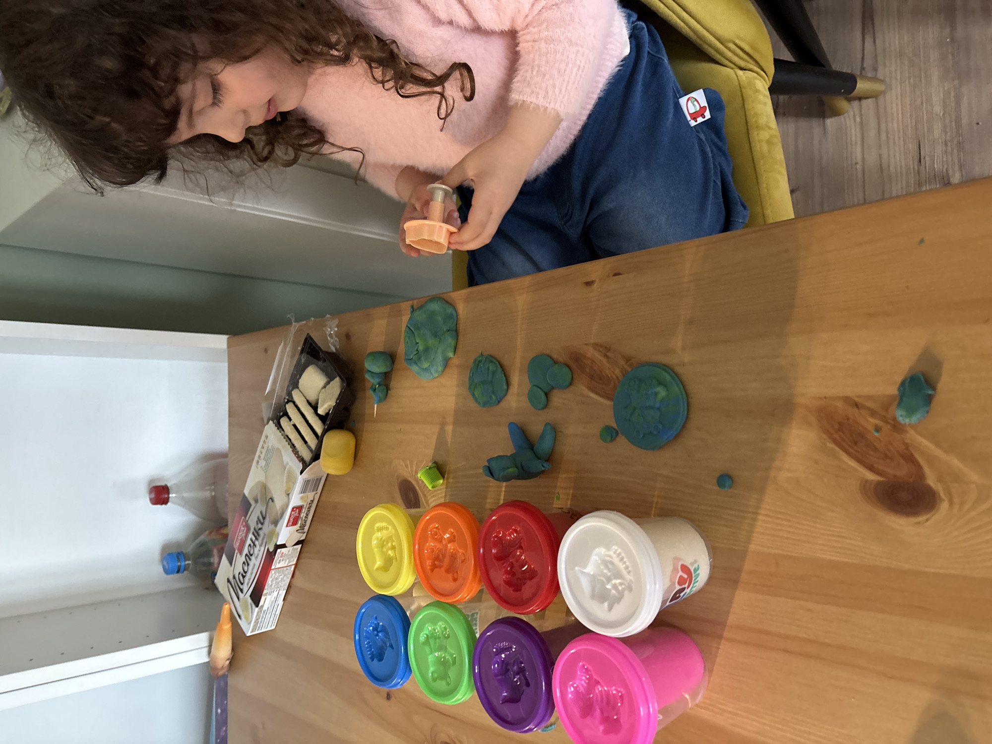 A child with curly brown hair and a pink sweater is seated at a wooden table, using a plastic cutter to make shapes in green and blue play dough. Eight containers of play dough in various colors (yellow, orange, red, white, blue, green, purple, and pink) are arranged on the table, along with scattered pieces of play dough. A package of "Maslenki" wafers and two plastic bottles are visible on the left side of the table near a white cabinet. The child is sitting on a yellow chair with a car sticker. The location appears to be a home interior, possibly in Sofia, Bulgaria, due to the "Maslenki" wafer brand, which is common in Bulgaria.
