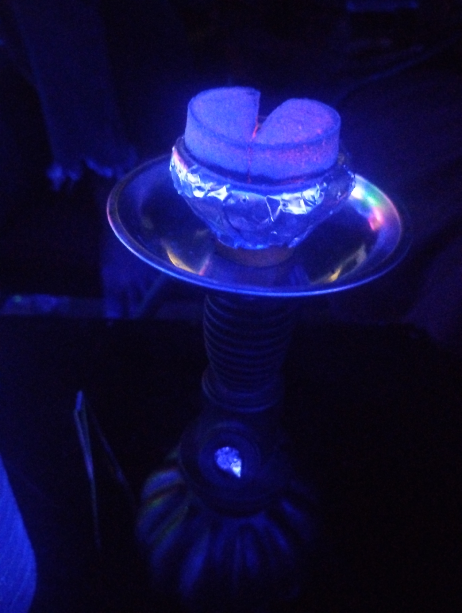 A hookah is shown in a dimly lit environment with blue light. The hookah features a silver tray on top, on which rests a layer of aluminum foil and two pieces of charcoal. The base of the hookah is bulbous with a visible light. The surrounding environment is dark.