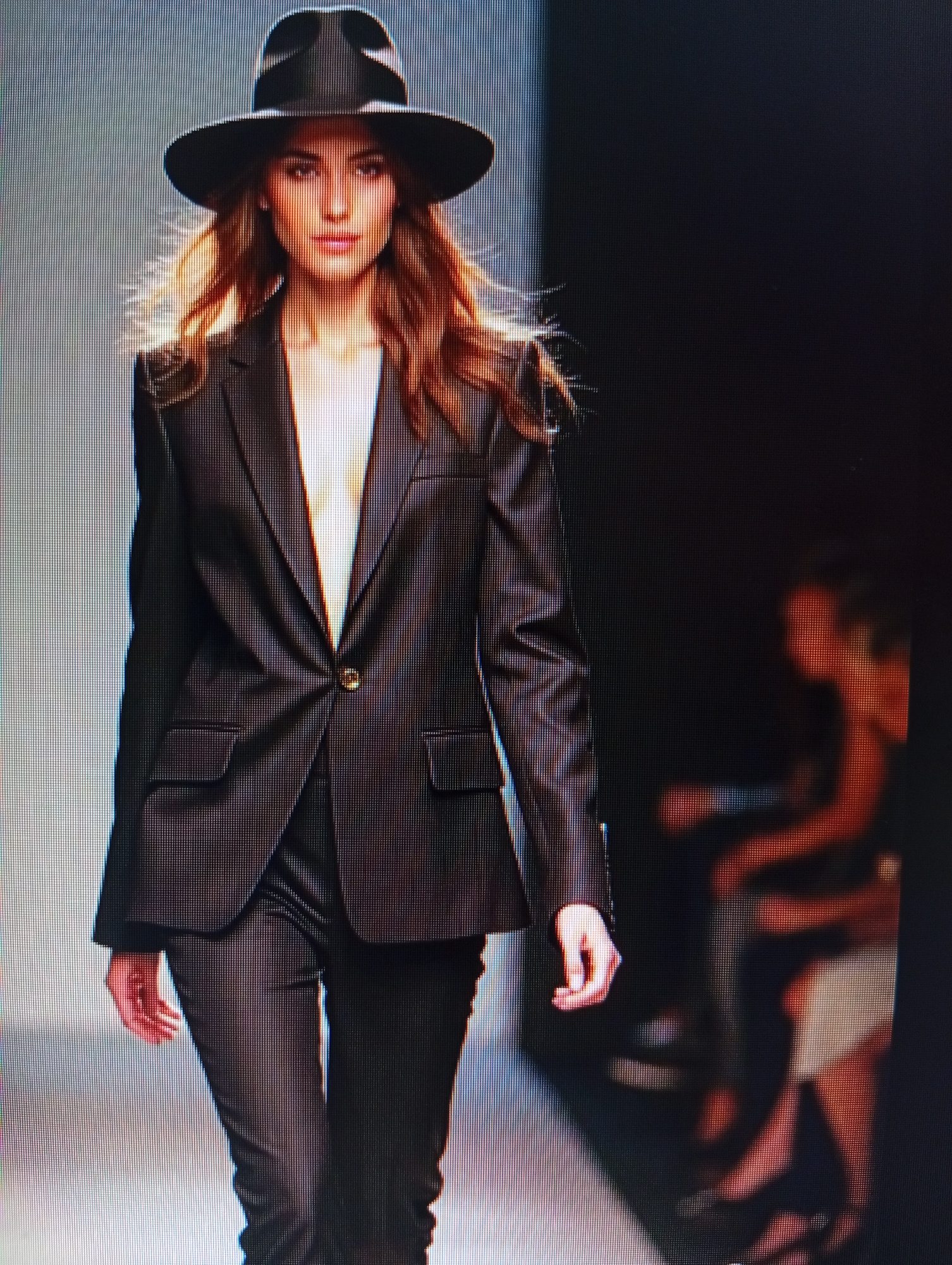 A fashion model walks down a runway wearing a black fedora and a dark suit. The suit consists of a single-button blazer with lapels and two flap pockets, paired with matching trousers. The model has long, wavy brown hair and a light complexion. A partially blurred figure is visible in the background to the right, appearing to be seated. The lighting is dramatic, with the model illuminated from the front and the background dark. The image appears to be a photograph of a screen, with visible pixelation and scan lines. The location is Unknown, Unknown.