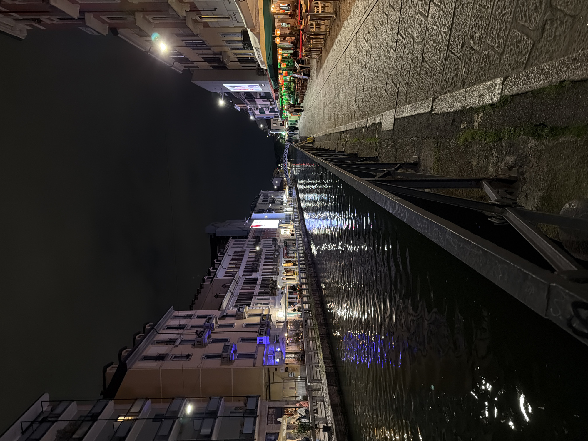 This media file captures a vibrant night scene along a canal in Milan, Italy, likely within the Navigli district. The image, oriented vertically, depicts a lively horizontal landscape.
On the right side of the frame, a textured, paved pedestrian walkway runs alongside a dark metal railing that borders the canal. The canal's water, dark and rippled, reflects numerous artificial lights from the opposite bank, creating shimmering streaks of white and blue across its surface.
To the left of the canal, a continuous row of multi-story buildings stands illuminated against a dark night sky. The ground floors of these buildings host various establishments, primarily restaurants and cafes, indicated by awnings and outdoor seating areas furnished with tables and chairs. People are visible along the walkway and seated at these outdoor venues, suggesting a bustling and social atmosphere.
Notable details include the characteristic European architecture of the buildings and the vibrant illumination that defines the scene. The time of day is clearly night. A green "OPEN" sign is visible on an awning of one of the establishments, but other signs are not legible. The scene conveys a lively urban environment, typical of a popular dining and leisure district after dark.