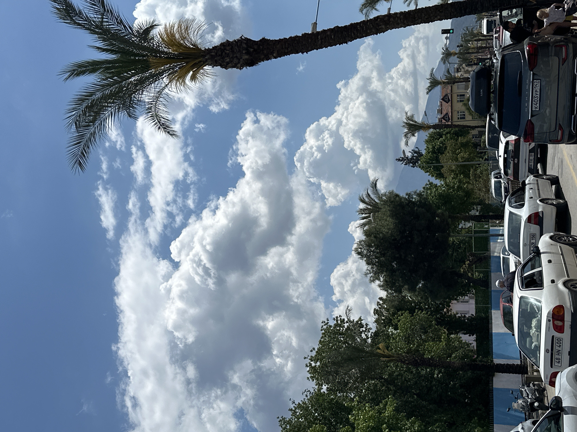 The image shows a street scene in Fethiye, Turkey. The sky is blue with fluffy white clouds. Palm trees are visible on both sides of the street. On the right side of the image, cars are parked along the street, including a dark gray SUV (license plate K403YP147). The front of the line of cars consist of 3 white cars. The parked vehicles create a slight obstruction in the background, where buildings and a mountain range are partially visible. A traffic light can be seen in the distance. The lighting conditions suggest it is daytime, with sunlight illuminating the scene.
