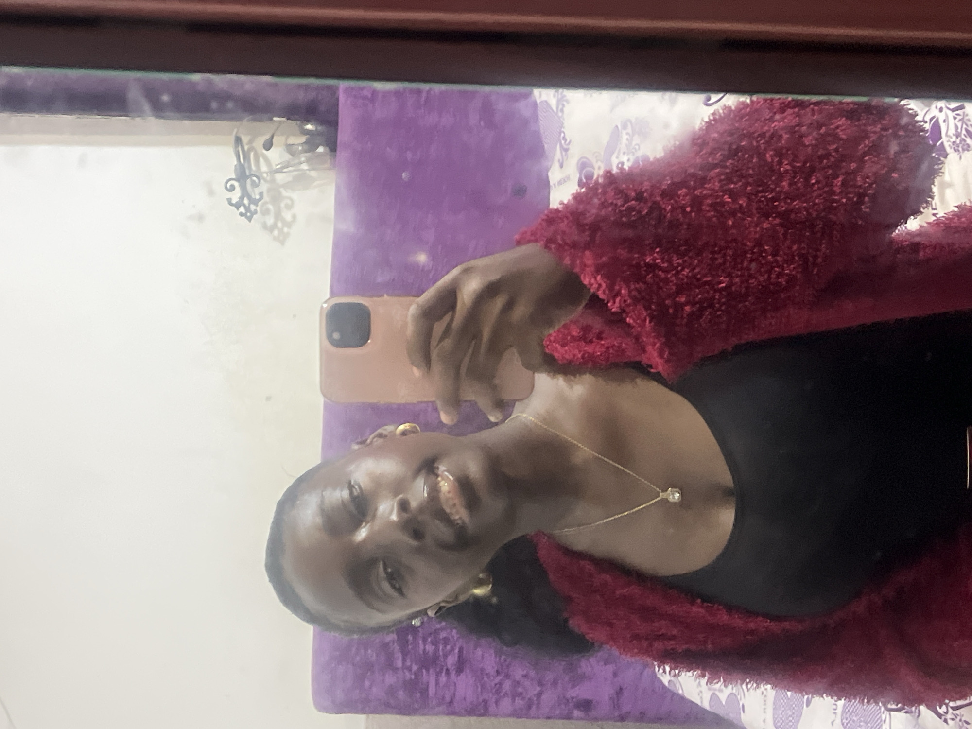 A person is taking a selfie in front of a mirror. The person is wearing a black top and a red fluffy cardigan. The person is holding a rose-colored phone. They are also wearing a gold necklace and gold hoop earrings. A purple cushion is behind the person. The location is unclear, with no details pointing towards Abuja, Nigeria.