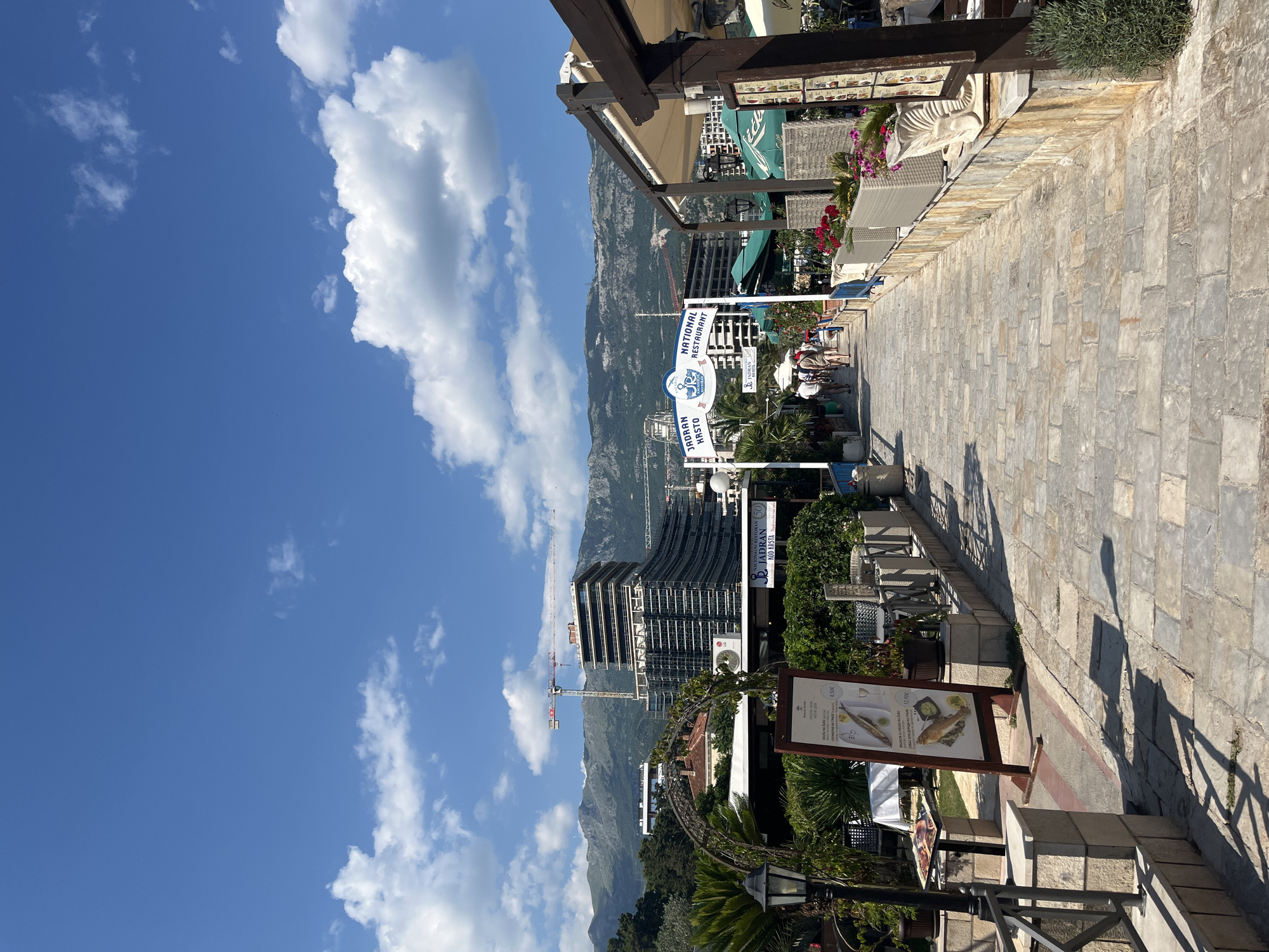 The image depicts a sunny day in Budva, Montenegro, focusing on a pedestrian walkway leading towards a national restaurant named "Jadran Krsto". The walkway is paved with stone tiles and lined with outdoor seating areas of restaurants on the right, featuring tables, chairs, and potted plants with flowers. On the left, there is a verdant area with palm trees, foliage, and a decorative archway, also with outdoor seating.
In the background, a large building complex is under construction, with cranes visible above it. Beyond the construction site, a mountain range forms a scenic backdrop under a bright blue sky with scattered white clouds. Several people are walking along the path, some appearing to be tourists. Signage for "Jadran Krsto National Restaurant" is prominently displayed. The scene suggests a lively coastal town during daytime.