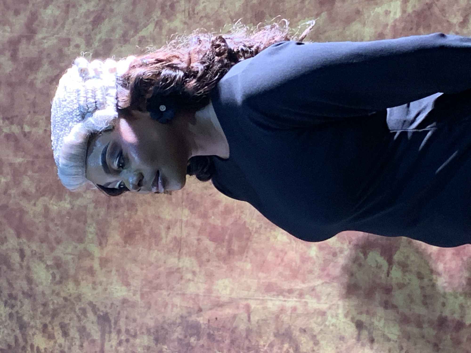 A woman wearing a judicial wig and a black dress stands in front of a mottled brown and orange backdrop. The woman is facing to the left, her face partially obscured. She has curly brown hair peeking out from beneath the wig, and is wearing a dark floral earring. The lighting casts a shadow on the right side of her face and highlights her cheekbones. The backdrop appears to be a textured fabric, possibly a stage backdrop. There is no indication of the city of Jos, Nigeria in the image.