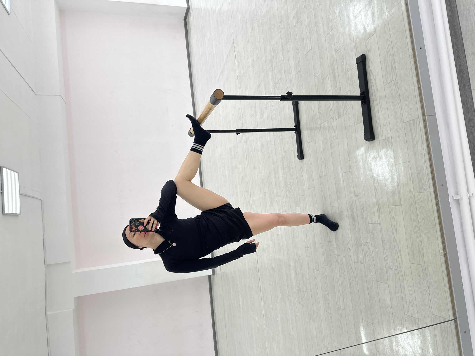 The image shows a person in a dance studio. The person is wearing a black long-sleeved shirt, black shorts, and black socks with white stripes, and appears to be a ballerina doing a ballet pose with one leg extended and resting on a ballet barre. The person is holding a cell phone up in front of their face, as if taking a selfie. The ballet barre is black with a wooden barre. The studio has a white wall and a light-colored wooden floor with a mirror on the right. It's unknown if this photograph was taken in Votkinsk, Russia.
