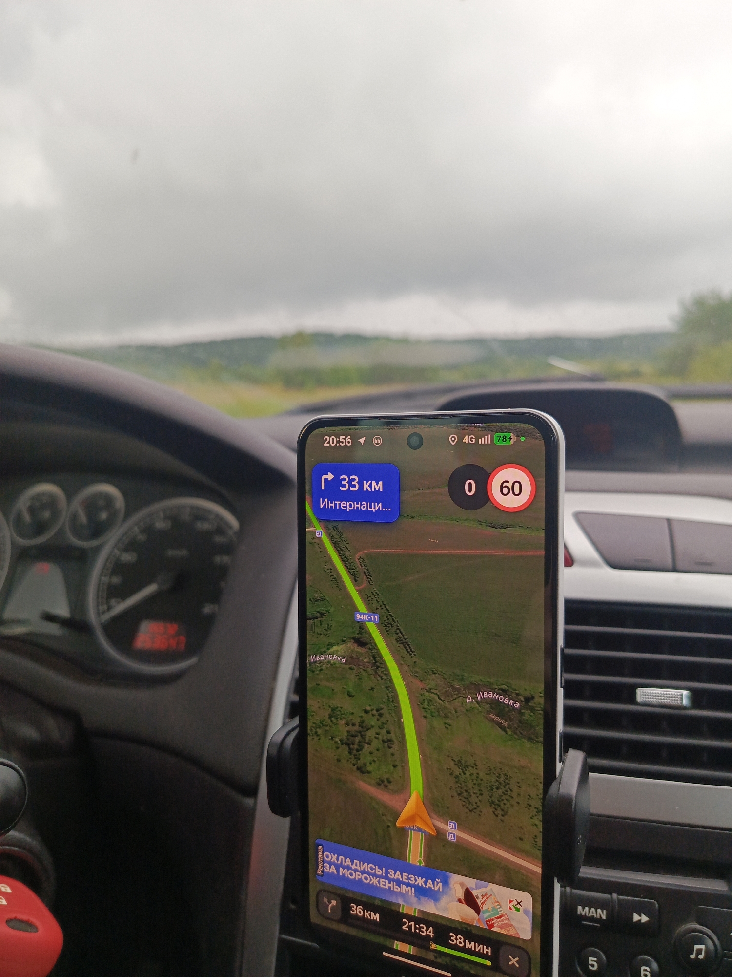 The image shows a car's dashboard with a smartphone mounted and displaying a navigation app. The phone shows a route with 33 km remaining to "Internatsiona...". The time displayed is 20:56, and the speed limit is shown as 60 km/h. The phone also displays "4G" signal strength. The car's instrument cluster is visible to the left, with a speedometer reading unclear but appearing to be around 150-160. The environment outside the car appears to be rural, with a cloudy sky and green landscape visible through the windshield. The car is likely in motion, given the navigation display and the speedometer reading. The location is specified as Makarovo, Russia, although this is not visually confirmed within the image itself.