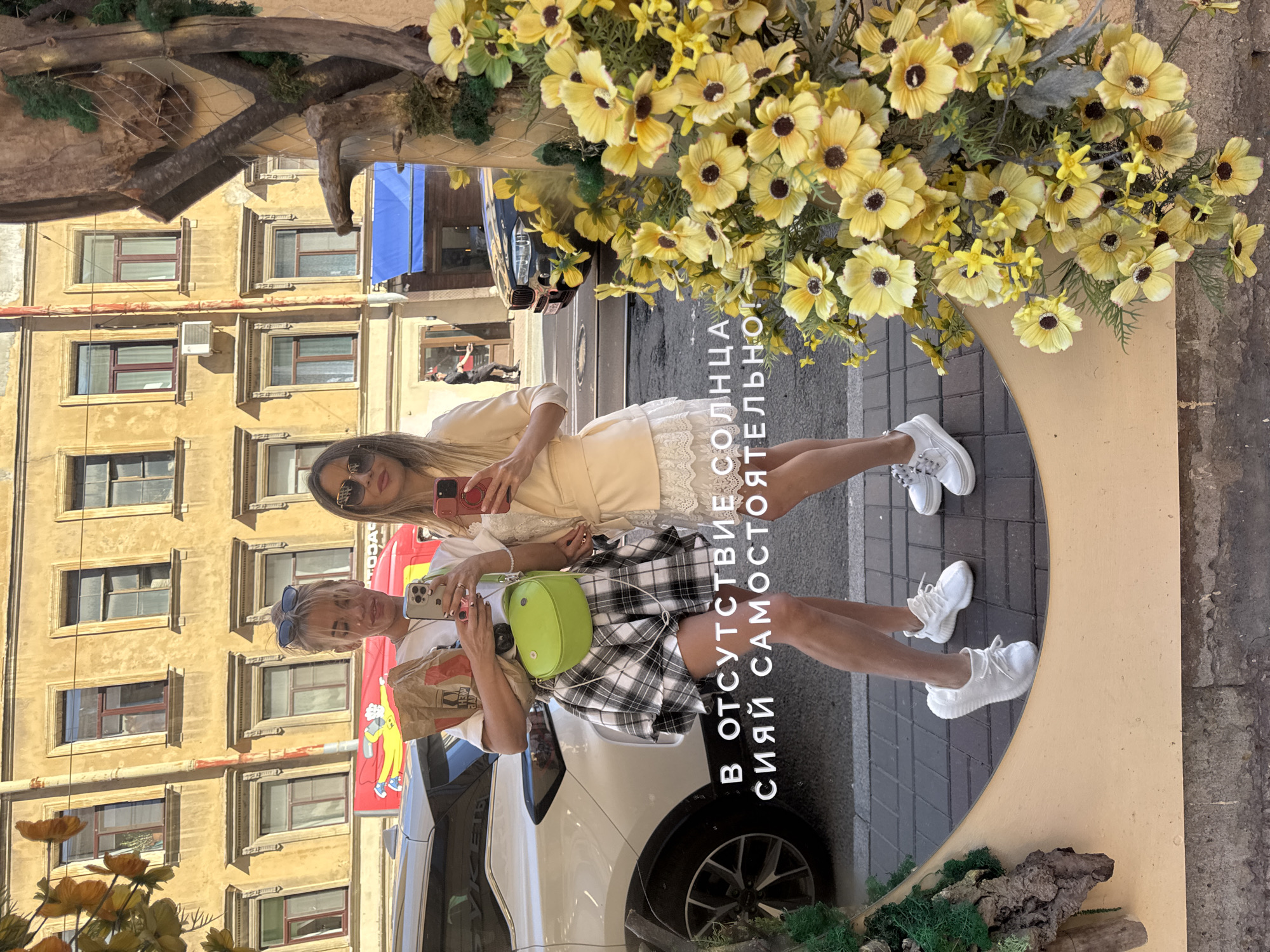 In Saint Petersburg, Russia, two women are captured in a mirror reflection, posing in front of a building with yellow facades and multiple windows. The woman on the left, with blonde hair, wears a white t-shirt, a black and white checkered skirt, and white sneakers. She holds a paper bag and a smartphone. Her green cross-body bag is slung over her shoulder. The woman on the right, with blonde hair and sunglasses, is dressed in a light-colored blazer over a white lace dress and white sneakers. She holds a smartphone. Their reflections are superimposed with Russian text. The foreground of the mirror is adorned with a large arrangement of yellow artificial flowers. In the background, reflected in the mirror, a white car is parked next to a building, and further down the street, a black car is visible. The scene suggests an outdoor urban setting, likely a street or sidewalk, on a sunny day.