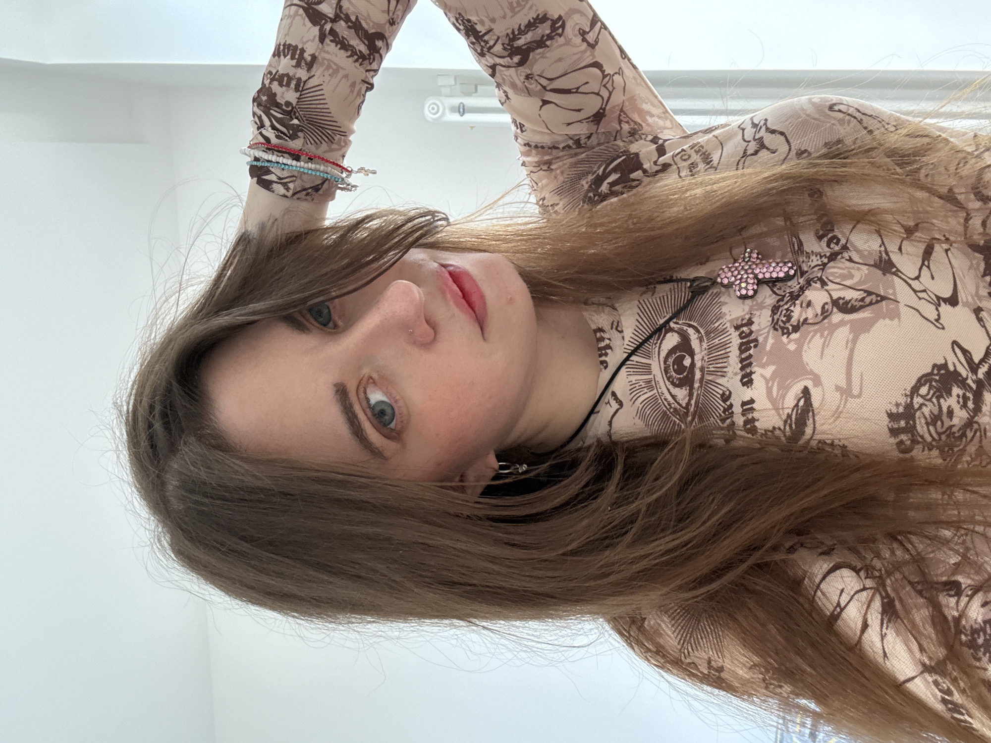 A young woman with light brown hair, fair skin, and light-colored eyes is facing the camera. She is wearing a beige, long-sleeved top with brown drawings of angels, eyes, and text. She has a nose piercing, small hoop earrings, and a black cord necklace with a pink bedazzled cross pendant. Multiple beaded bracelets are on her wrist. The background consists of a white wall and a glimpse of a light fixture. It is impossible to determine the location is in Moscow, Russia, from this image.