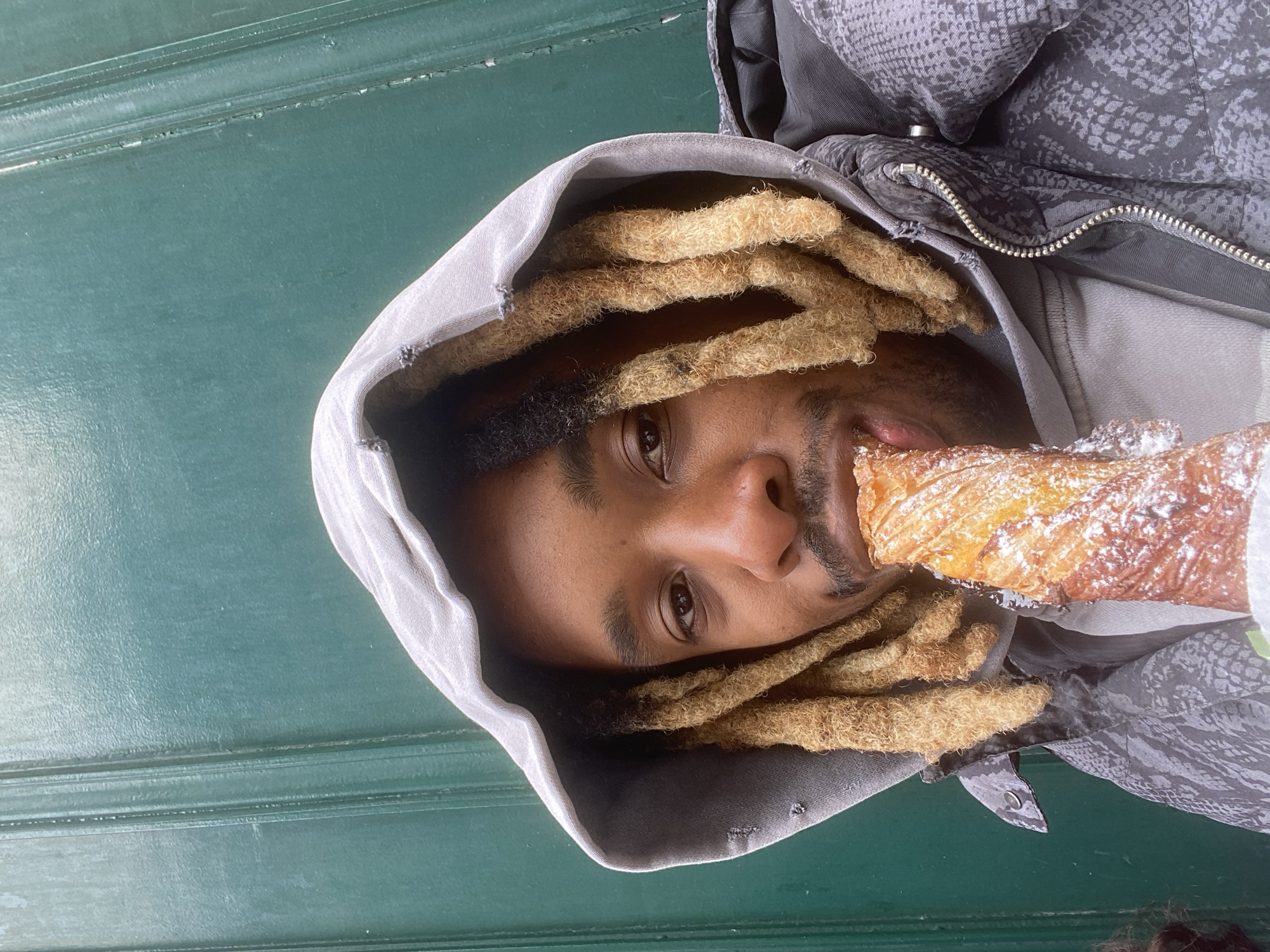 A man wearing a hooded sweatshirt and with dreadlocks, is eating a pastry covered in powdered sugar in front of a dark green paneled surface, possibly a door or wall. The setting is implied to be the city of Grenoble, France. The man is turned sideways to the camera, with the pastry near his mouth. He appears to be indoors or next to a building.