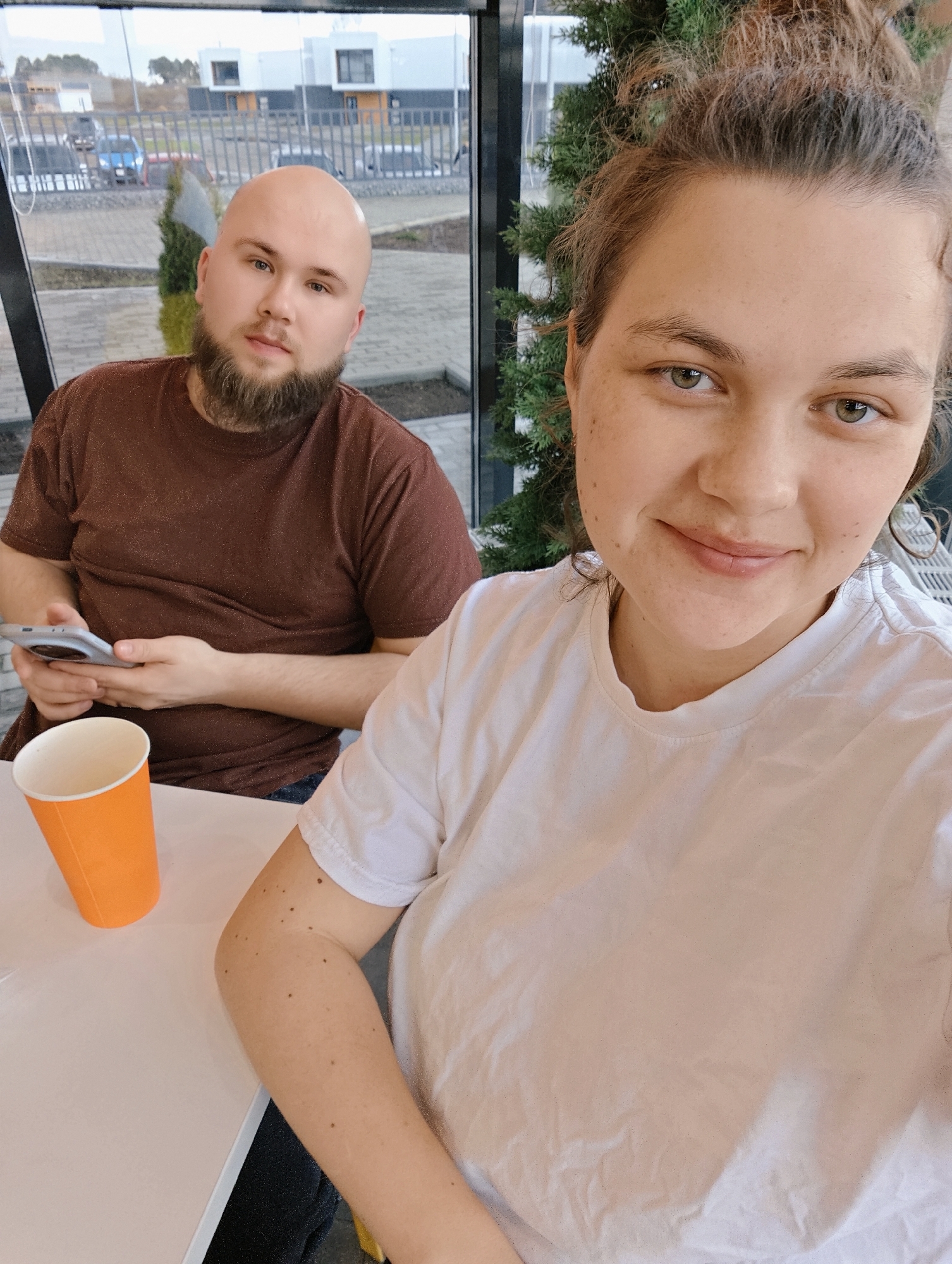 The image features two individuals indoors, possibly in a cafe or a modern building with large windows, set in Votkinsk, Russia. The woman on the right, in the foreground, is taking a selfie, smiling gently at the camera. She has light brown hair pulled back, visible freckles on her face and arm, and is wearing a plain white t-shirt. On the left, in the background, a bald man with a full brown beard and moustache is looking towards the camera with a neutral expression. He is wearing a brown t-shirt and holding a silver smartphone.
An orange paper cup rests on the white table in the foreground. Between the subjects, near the window, is a small, dark green artificial plant. Through the large window, an outdoor scene is visible: modern white and dark grey buildings, a paved area, a dark fence, and several indistinct vehicles. The lighting suggests it is daytime with an overcast sky, creating soft, diffused light. The overall mood is relaxed and casual. No visible text is present in the image.