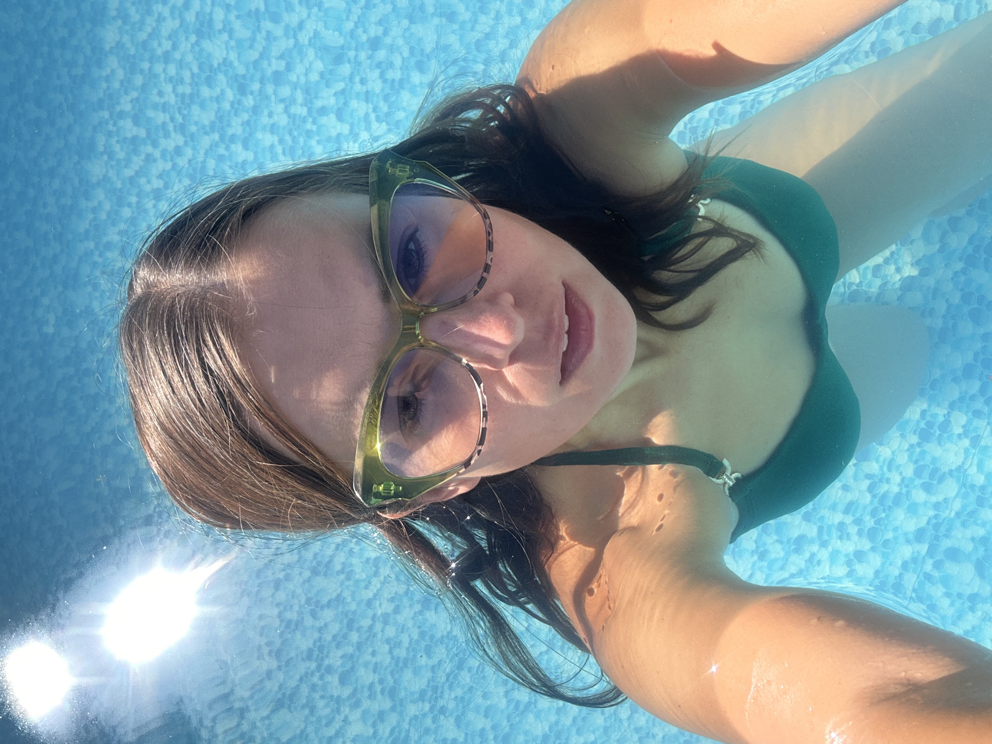 A woman is underwater in a swimming pool, taking a selfie. She is wearing a green bikini and stylish sunglasses. Her hair is wet and flowing around her face. The water is clear and blue, with the sunlight creating bright reflections on the surface. The bottom of the pool is tiled with small, light blue and white squares. The lighting suggests it is daytime, with the sun high in the sky.