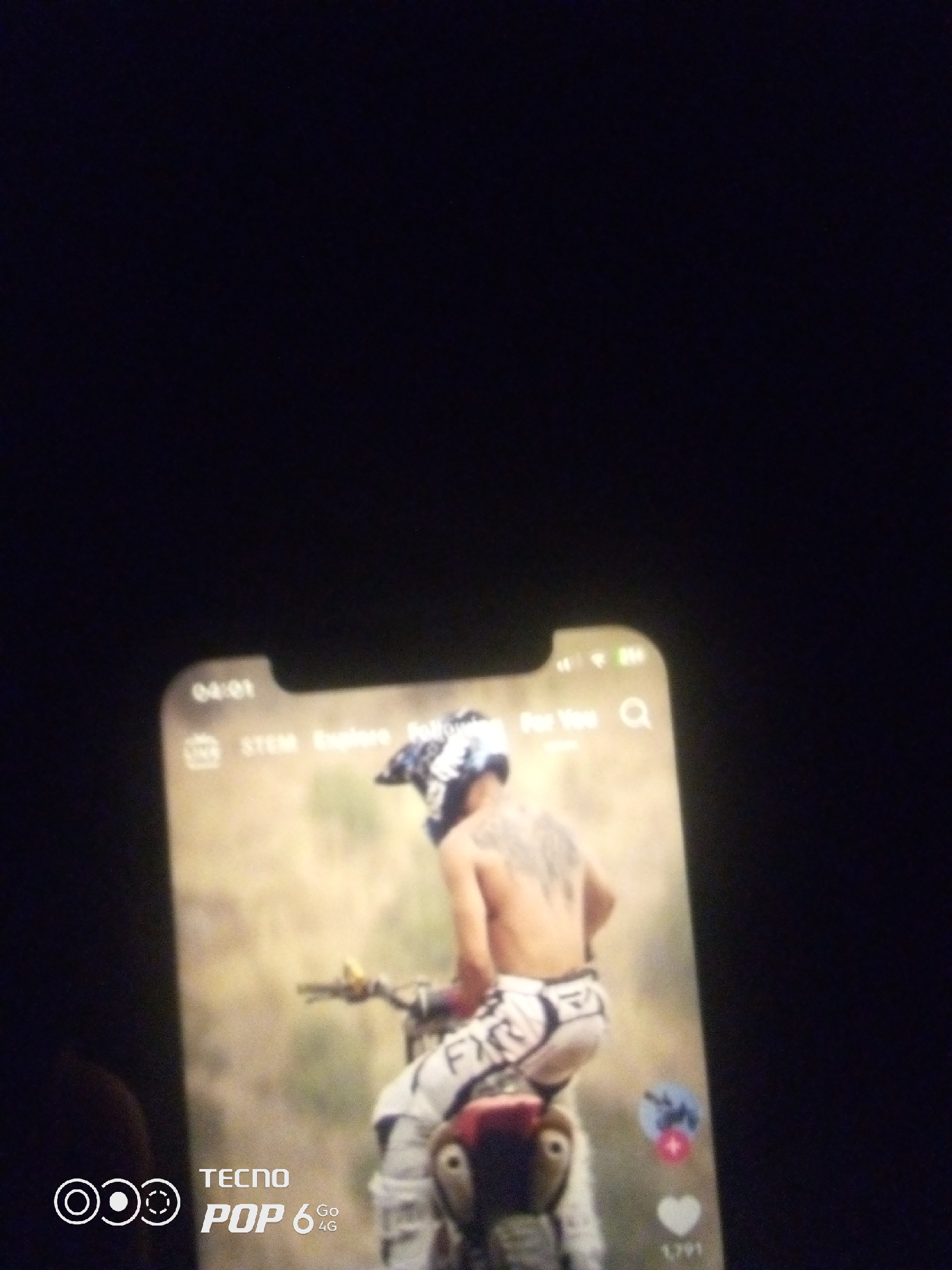 The image shows a mobile phone screen displaying a video of a shirtless person with tattoos riding a dirt bike. The person is wearing a helmet and racing pants. The background of the video appears to be an outdoor, possibly rural, setting with dry vegetation. The phone itself is a TECNO POP 6 4G. The phone's interface elements are visible, including the time "04:01", icons for "STEM Explore", "For You", and a search icon. Social media interaction icons are also visible on the right side of the video, including a profile picture and a heart icon with a number. The image does not contain any information that would indicate a location in the city of Howayi, Nigeria, or any specific geographical details.