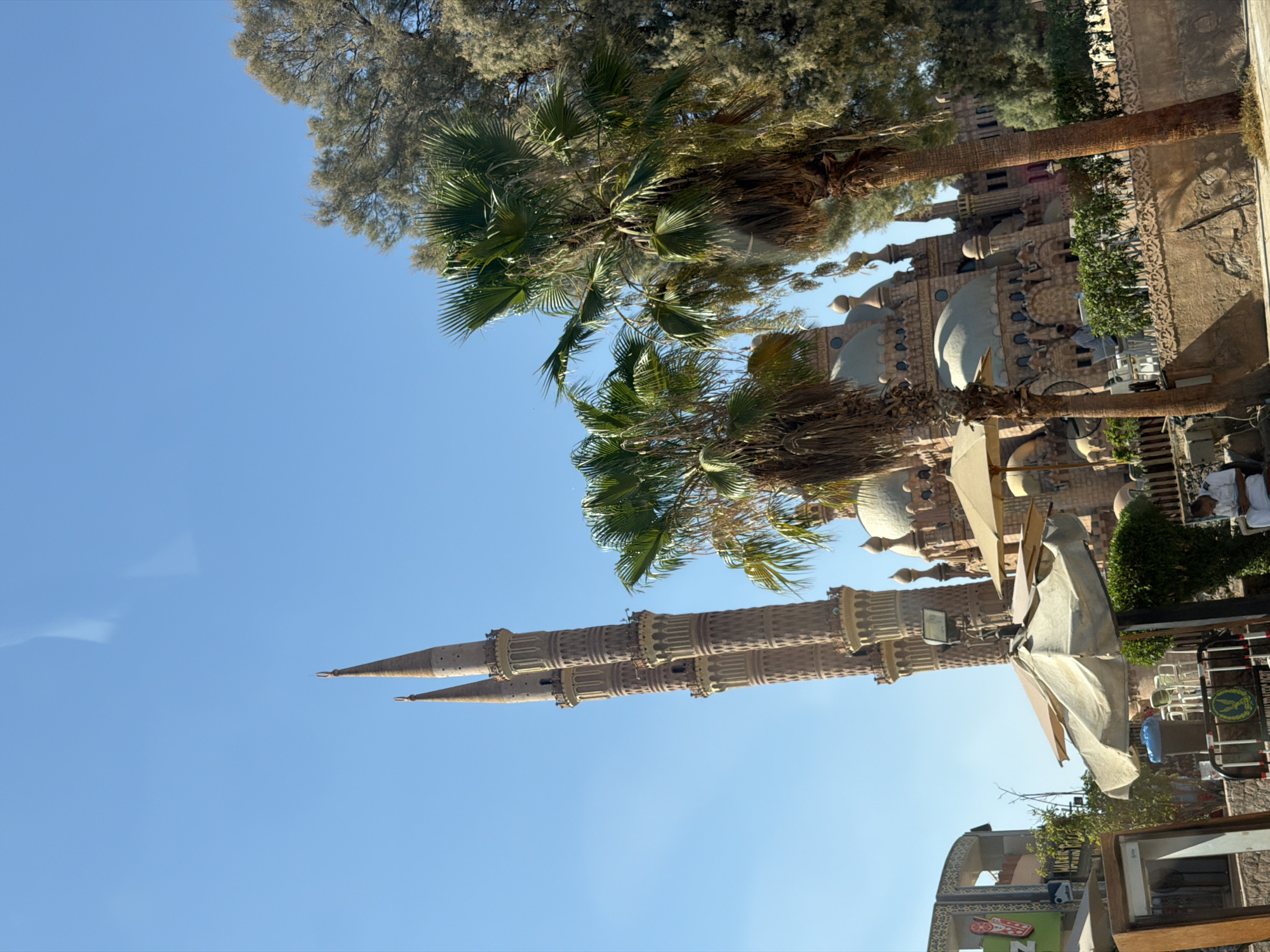 The image captures an outdoor scene dominated by the Al Sahaba Mosque in Sharm El Sheikh, Egypt, on a clear day. Two tall, intricately designed minarets with multiple tiers rise prominently against a bright blue sky. In the foreground, several palm trees with lush green fronds partially obscure the view of the mosque's domes and architectural details.
In the lower portion of the image, there are elements of a public space or seating area. White and beige umbrellas are set up, providing shade. White metal chairs and tables are visible, suggesting an outdoor cafe or resting area. A man in a white uniform sits on a bench to the right, looking down. A decorative stone wall with ornate carvings runs along the foreground. To the left, a building facade with signage is partially visible. The overall impression is a blend of religious architecture and a recreational outdoor environment in a warm, sunny climate.