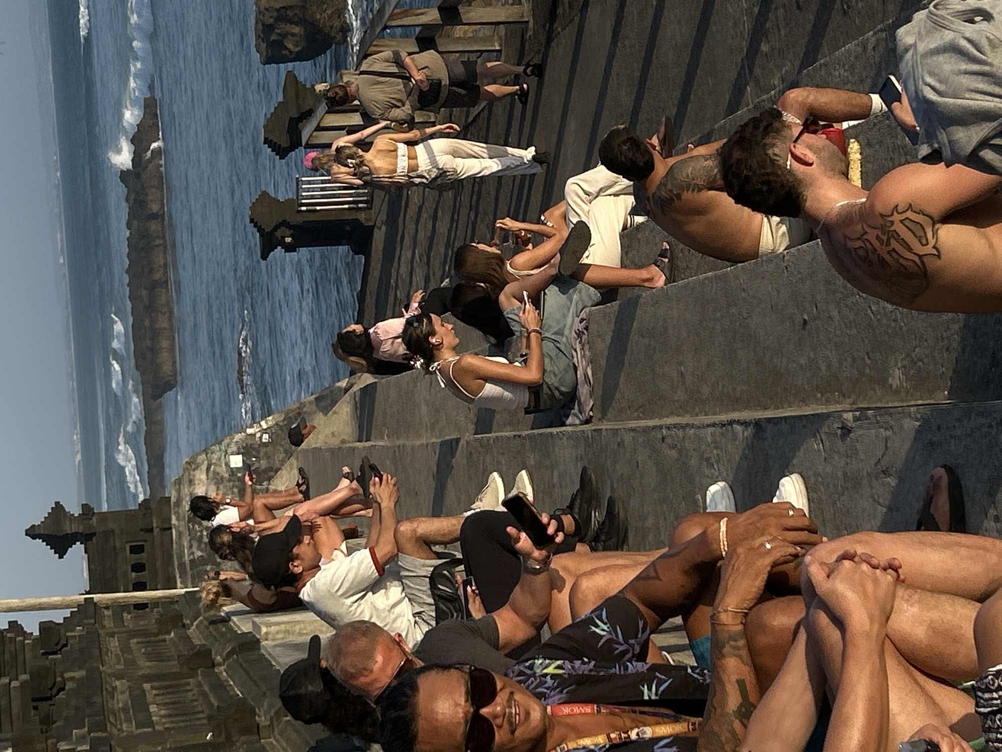 The image depicts a group of people gathered on stone steps overlooking the ocean in North Kuta, Indonesia, likely near the Luhur Uluwatu Temple. The setting appears to be late afternoon, with the sun casting long shadows. Several individuals are seated, engaged with their mobile phones or observing the ocean.
In the foreground, a man with sunglasses and a floral shirt is looking towards the camera, with visible tattoos on his arms. Next to him, another person is seated, also with tattoos. Further back, a group of people are seated on the steps, some of whom are looking at their phones.
Beyond the steps, the ocean stretches out, with waves crashing against dark rock formations. The architecture of a traditional Indonesian temple is visible on the left side of the frame, adding to the cultural context of the location. A pathway runs along the edge of the cliff, with more people walking and observing the scenery. The overall atmosphere suggests a relaxed and tourist-oriented environment, where people are enjoying the coastal views and the ambiance of the area.