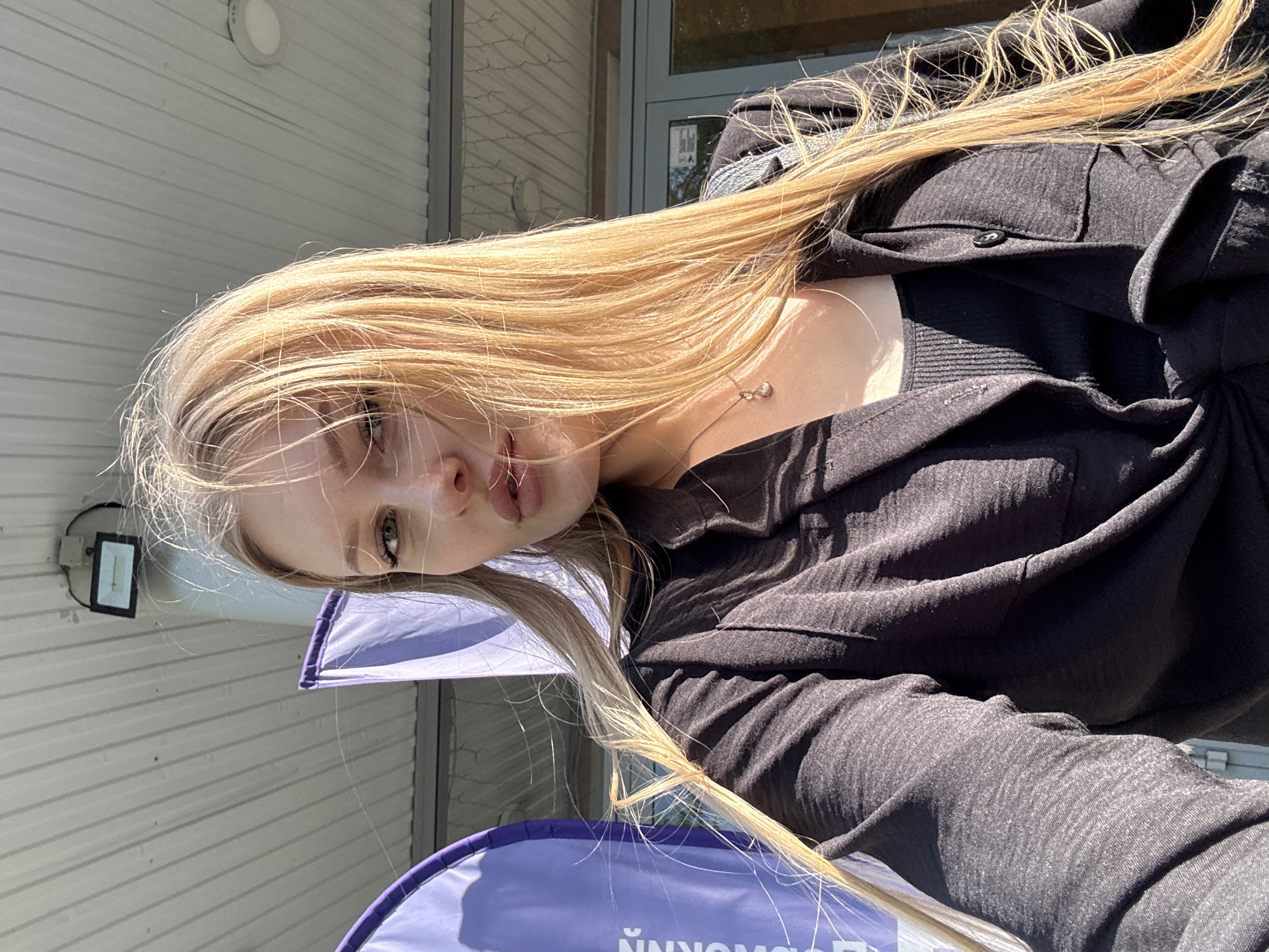A young, fair-skinned, light-haired woman is in the foreground, angled downward in a selfie. She is wearing a black top and has a silver necklace. A round, lavender-colored object with text in Cyrillic characters is visible below her. The background features a light-colored building with horizontal siding and a door. A small, rectangular light fixture is on the building. The location is likely Perm, Russia, based on the Cyrillic text.