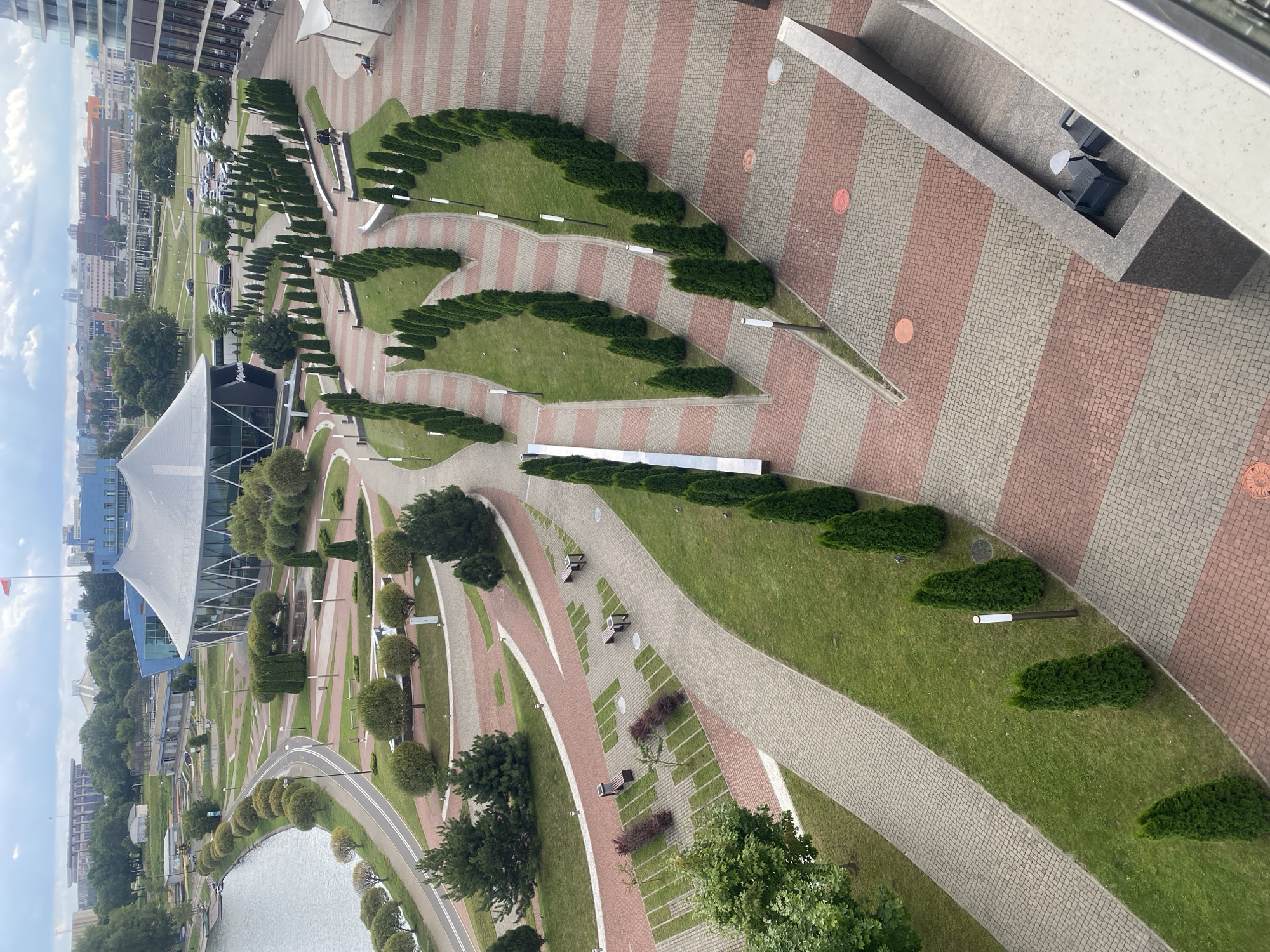 This is an aerial, full-frame, daylight, outdoor shot of a modern architectural complex situated on the waterfront in Minsk, Belarus, likely near the Minsk Arena. The scene is dominated by a large, tent-like building with a silver roof and glass walls, possibly a sports or concert venue. Adjacent to it is a body of water, likely the Svislach River, with a pathway and bike lane running alongside.
The foreground features a paved plaza with a distinct pattern of red and gray interlocking pavers. This plaza is landscaped with rows of manicured evergreen trees, interspersed with grassy areas and modern lampposts. Several benches are strategically placed throughout the plaza, with two individuals seated on one. Further into the scene, the plaza transitions into a park-like area with more trees and winding walkways.
In the background, a cityscape unfolds with various modern buildings, including office towers and residential complexes, along with a highway and parked cars. The sky is overcast with some visible clouds. The overall impression is one of a well-designed urban recreational and entertainment area, blending modern architecture with natural landscaping.