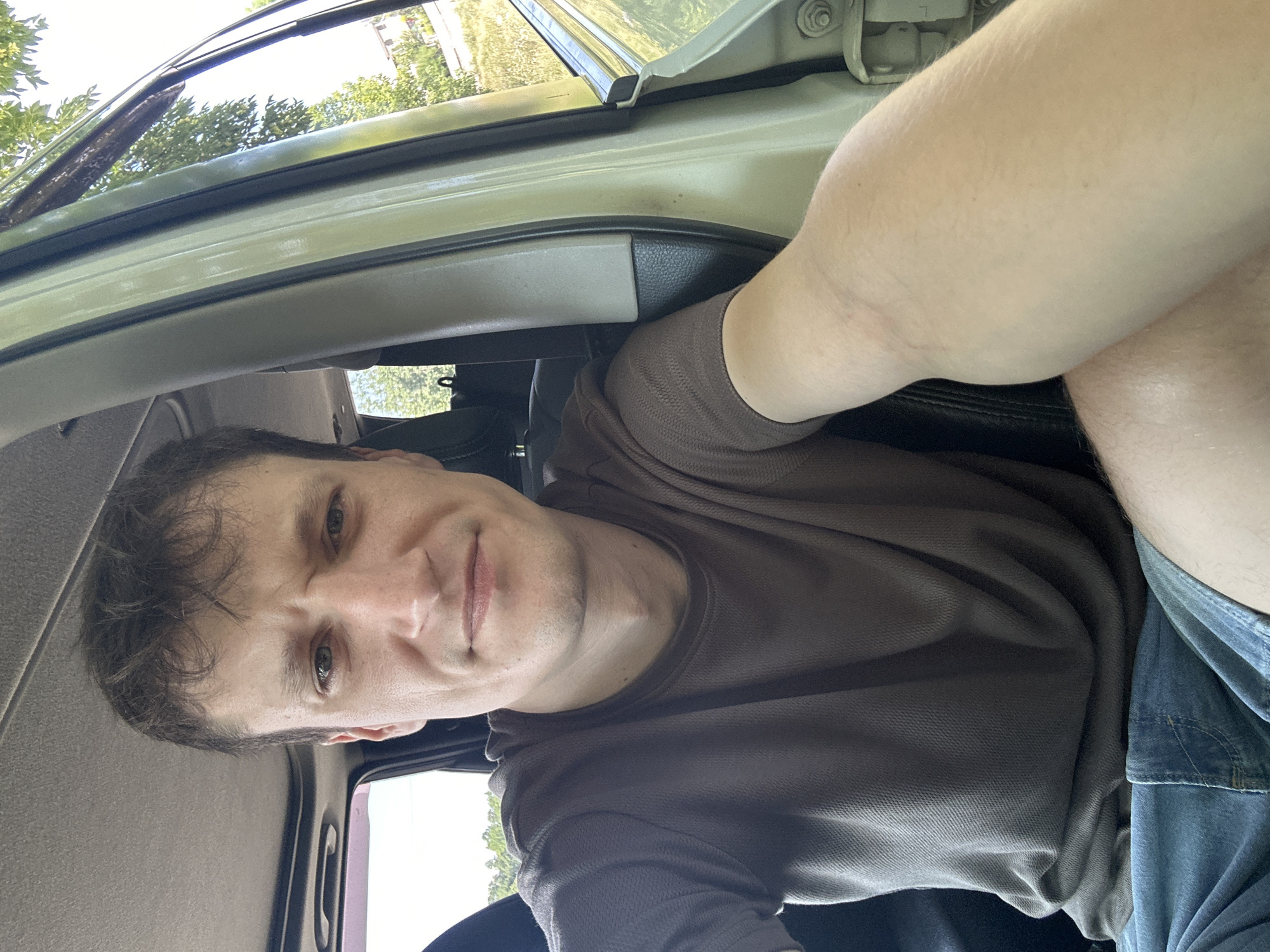 A man sits in the driver's seat of a car, taking a selfie. He is wearing a brown t-shirt and blue denim shorts. The interior of the car is visible, including the dashboard, door panel, and a sunroof. Outside the car, trees and a building can be seen. The scene appears to be in a rural or suburban area, possibly Makiv, Ukraine, given the context. The man's expression is neutral, and his arm is extended to capture the shot.