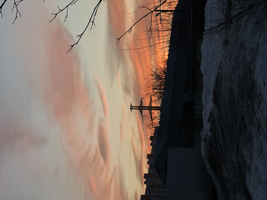 An outdoor scene captured at either dawn or dusk in the city of Khimki, Russia, displays a vibrant sky transitioning from pale blue and grey at the zenith to intense orange and red near the horizon, populated by scattered, elongated pink and orange clouds. The lower two-thirds of the image are dominated by silhouetted urban and industrial elements.
In the mid-ground, a prominent, dark-silhouetted electrical transmission tower (pylon) stands centrally, with its lattice structure visible against the brighter sky. To its left, a row of multi-story residential apartment buildings is partially visible, also in silhouette. Below these, a collection of darker, lower-profile industrial buildings or warehouses with various rooflines stretches across the frame. Bare tree branches, characteristic of deciduous trees in winter, are interspersed throughout the mid-ground, particularly along the right side and near the industrial structures.
The foreground consists of irregular piles of snow, appearing dirty and partially melted, covering the ground. Some sparse, bare branches protrude from these snowdrifts. On the far left of the foreground, a large, rectangular, light-colored object, possibly a section of a building or a container, is visible, cast primarily in shadow. No individuals or specific actions are discernible within the frame. The overall lighting suggests a low-light condition, with strong contrast between the luminous sky and the dark, silhouetted terrestrial features.