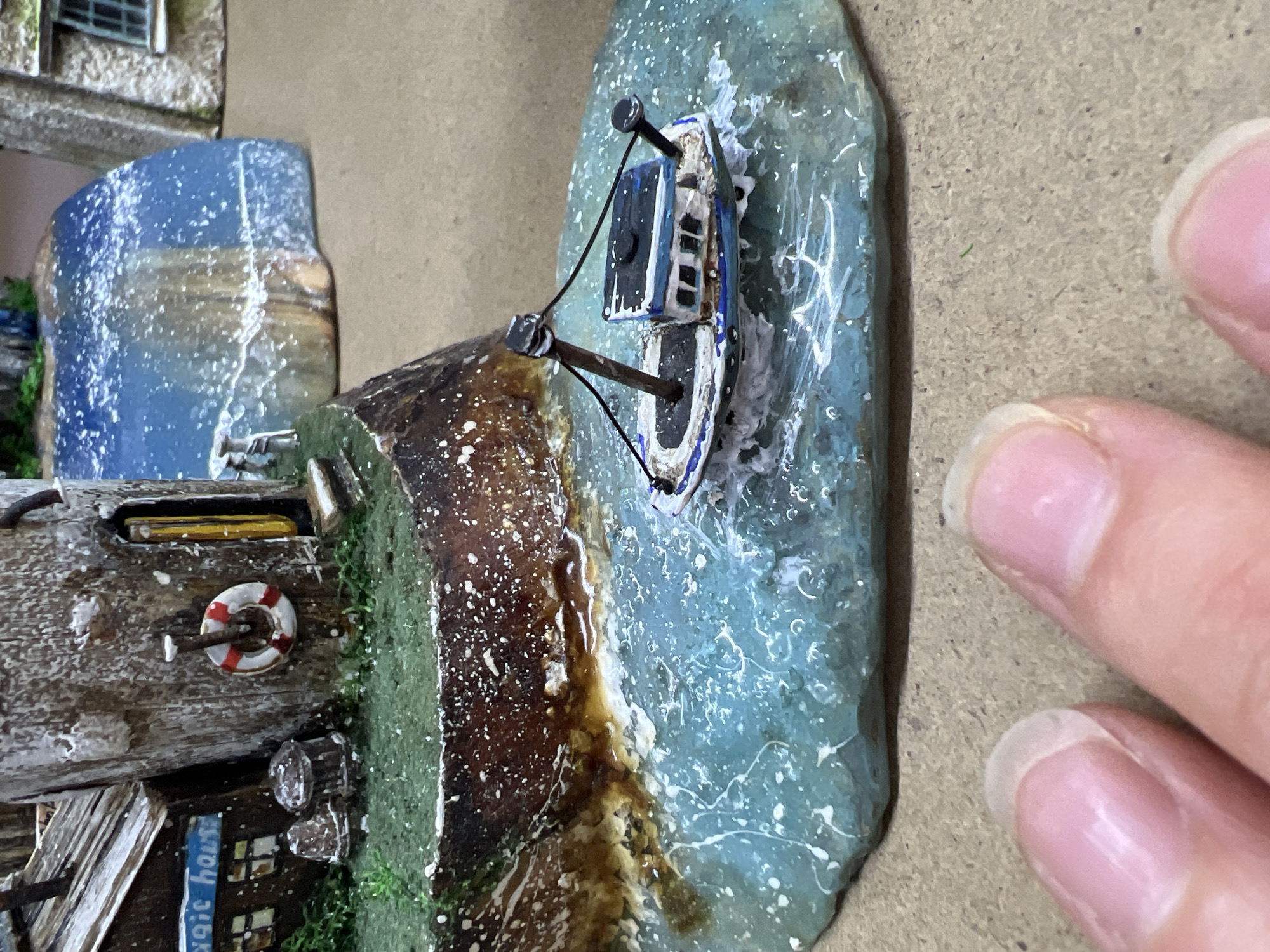 A miniature diorama depicts a coastal scene. A small boat is shown in the water, near a building with a life preserver attached to its wall. Part of a hand is visible on the right side of the image. The scene is likely a crafted art piece. There is no clear indication of its location being in the city of Chaikovsky, Russia.