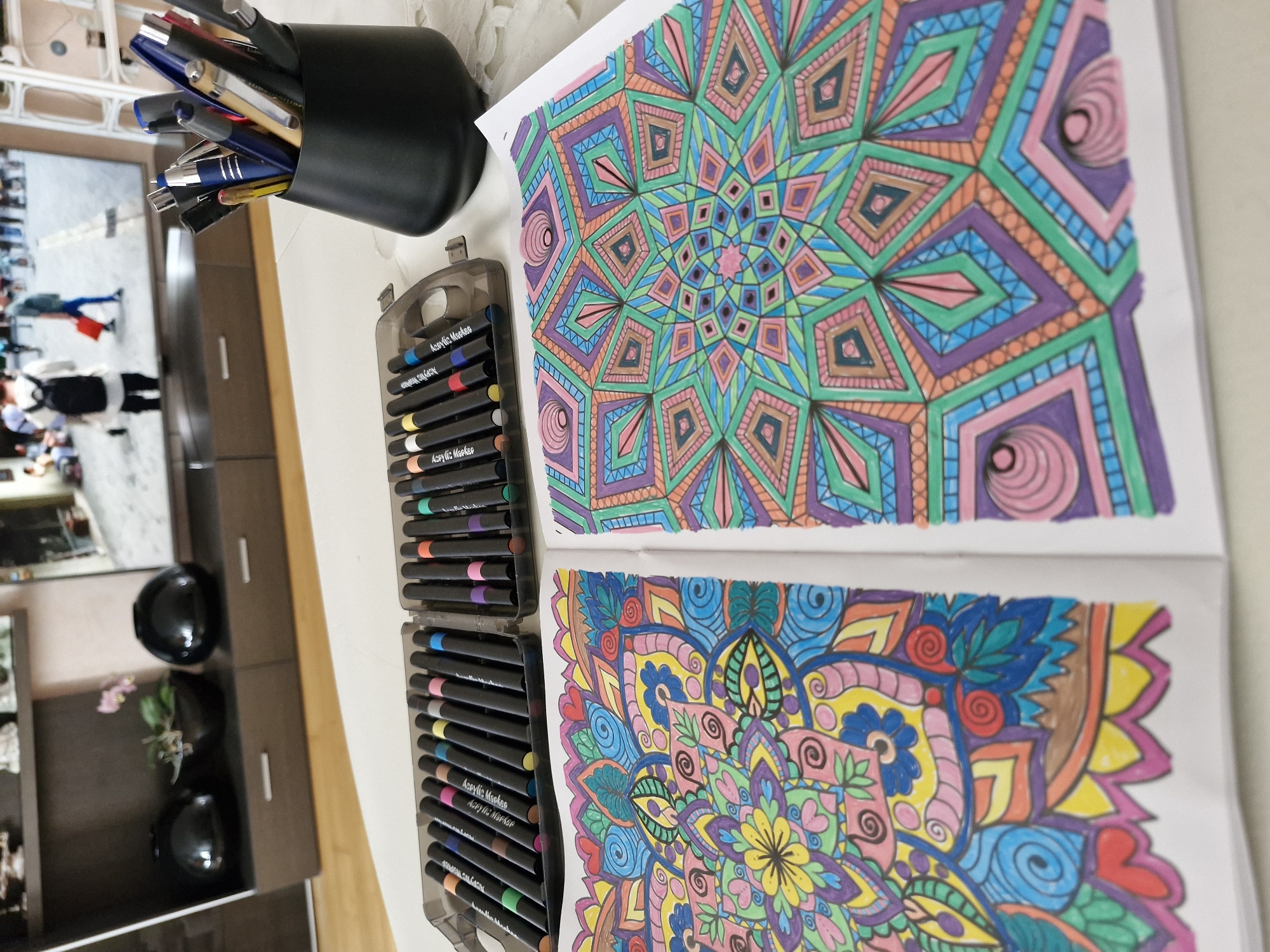 The image depicts a close-up view of an open coloring book featuring two intricate mandala designs. The left page showcases a mandala with organic, floral-like patterns in vibrant hues of pink, yellow, blue, green, and red. The right page displays a more geometric mandala with symmetrical star-like shapes rendered in shades of green, purple, pink, and orange. In the background, a black pen holder is filled with various pens and markers. To the left of the pen holder, a grey plastic case holds a set of black-barreled markers. Further in the background, a television screen displays a blurred image of people walking on a street, possibly in an outdoor setting. The coloring book and art supplies are placed on a white surface, which appears to be a table. The overall scene suggests a creative activity taking place indoors. The image does not provide any visual cues to suggest a specific location in the city of Bremen, Germany.