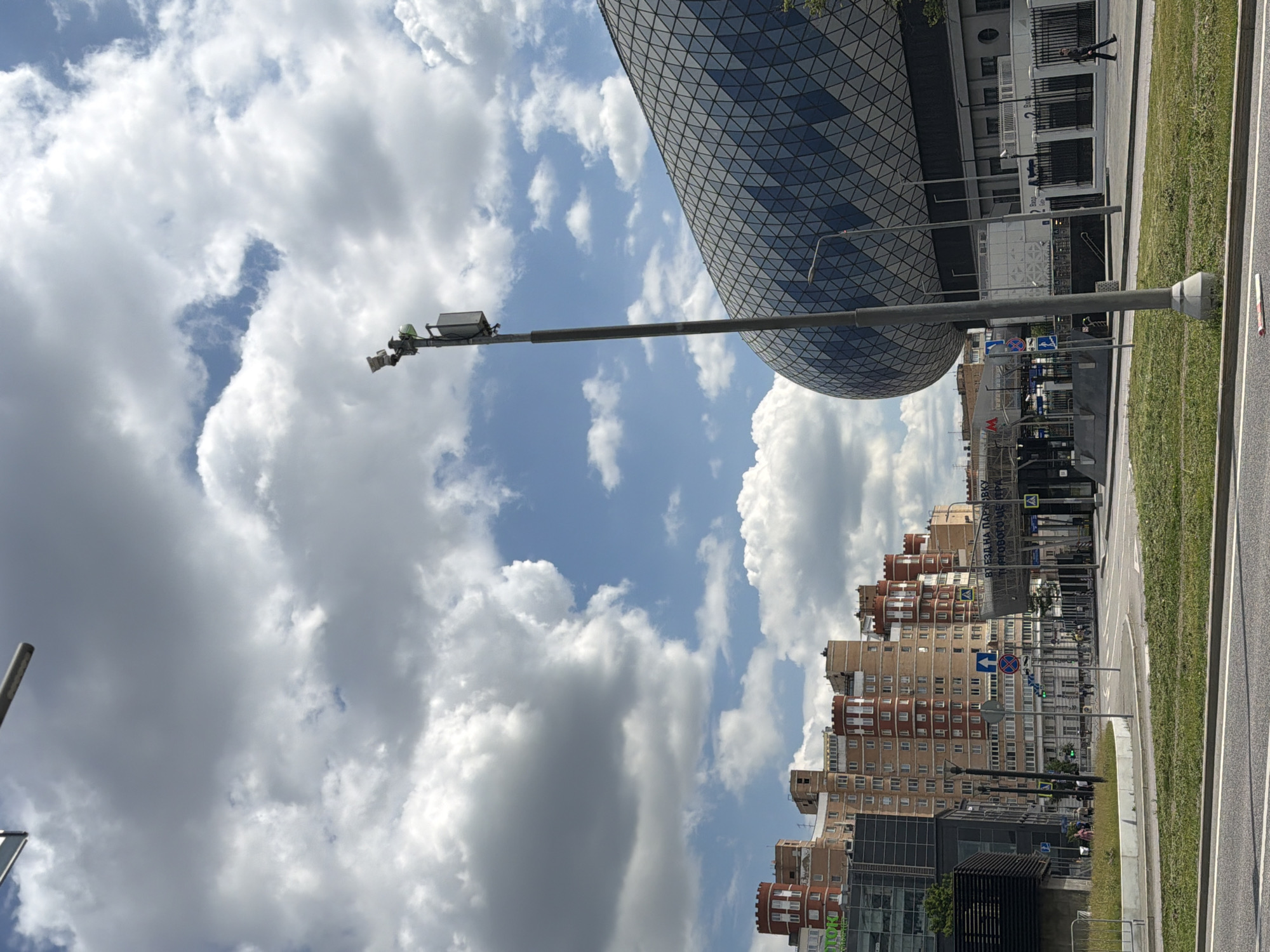 A street scene unfolds under a cloudy blue sky, featuring a prominent lamp post in the foreground. To the left, a series of multi-story buildings with a distinctive red-capped, turret-like architectural style line the horizon. In the mid-ground, a modern, curved building with a patterned facade, possibly a stadium or arena, dominates the right side of the frame. Signs are visible, including what appears to be a metro station entrance. A few individuals are present: one person walks along a sidewalk in the mid-ground, and a few more are seen near the buildings and the metro entrance. The environment is urban, with roads, sidewalks, and grassy areas.
