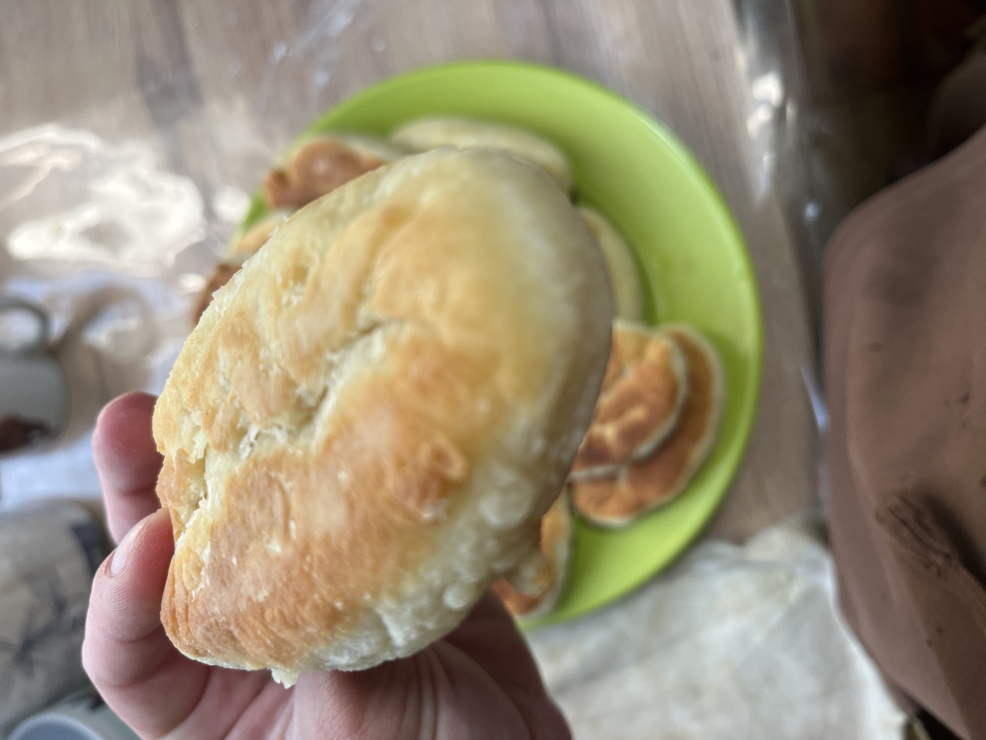 A person's hand holds a golden-brown pastry over a bright green plate piled with similar pastries. The pastries appear to be fried dough, possibly pirozhki, with a slightly puffed, uneven surface. In the background, out-of-focus mugs and what looks like a plastic bag are visible. The scene is set on a wooden table. There is no information about the location in Nizhny Tagil, Russia.
