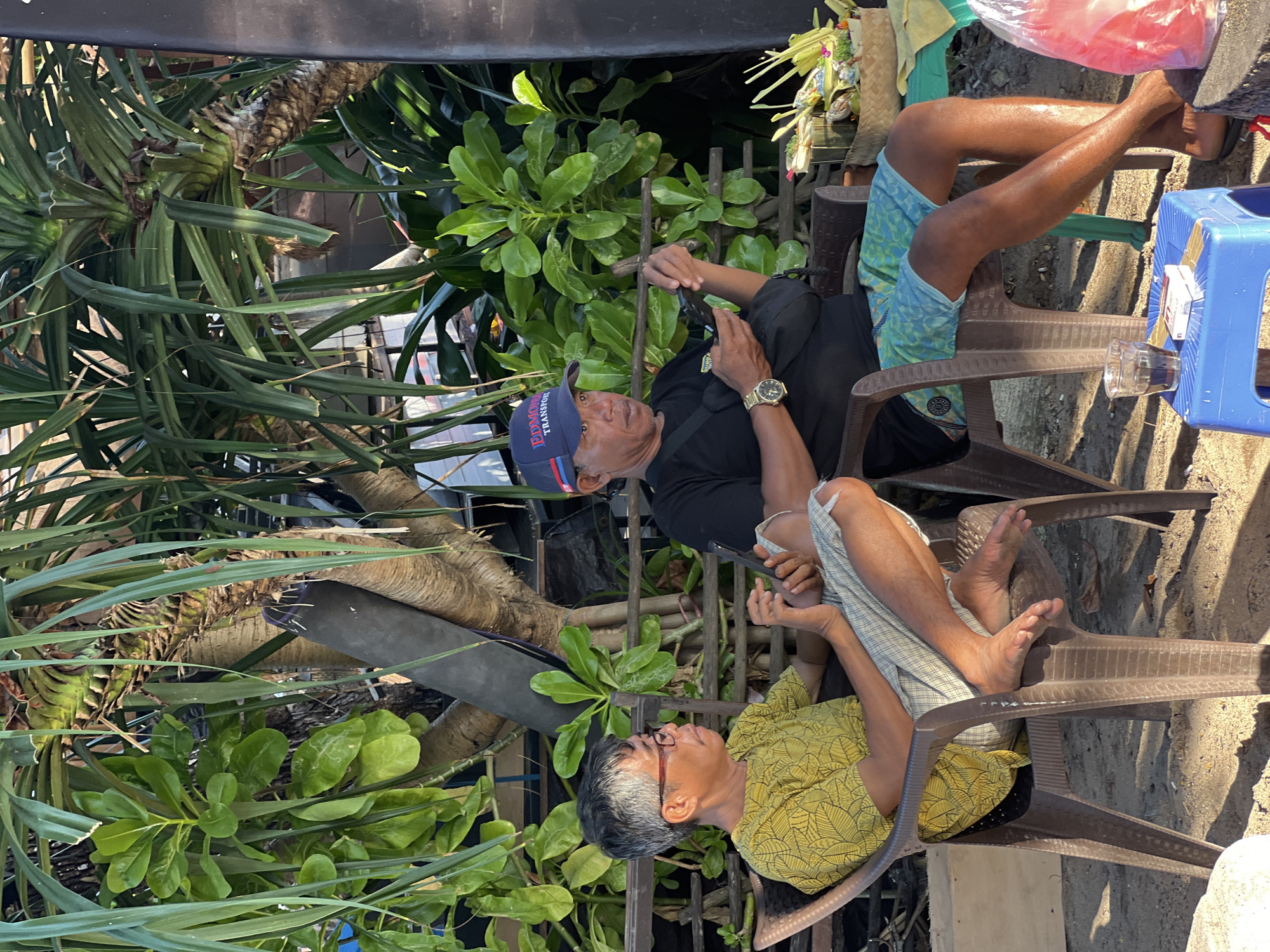 Two men are seated on plastic chairs outdoors, with lush green foliage and a rough-barked tree behind them. The man on the left, wearing glasses and a yellow patterned shirt, holds a mobile phone in his hands, looking down at it. The man on the right, wearing a blue baseball cap with "EDMON TRANSPORT" visible and a black t-shirt, also holds a mobile phone and looks towards it. He is wearing patterned blue shorts and a watch. In the foreground, a small blue plastic stool holds a glass of dark liquid, a pack of cigarettes, and some small, decorative items in woven baskets. The ground is sandy. The scene appears to be in Denpasar, Indonesia.