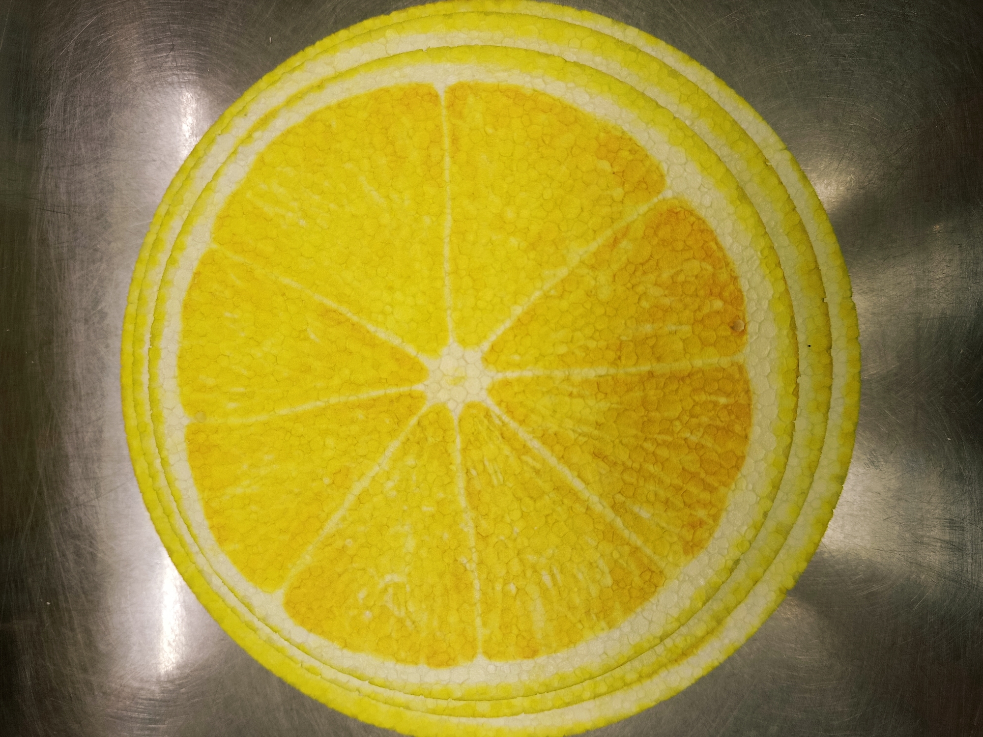 An overhead shot shows a circular, yellow and white lemon slice design printed on a mat. The mat is placed on a metallic, scratched surface. The lemon slice design features concentric rings representing the rind and pith, with segments emanating from the center. The texture of the printed design mimics the bumpy surface of a lemon. The metallic background has visible scratches and reflects light.