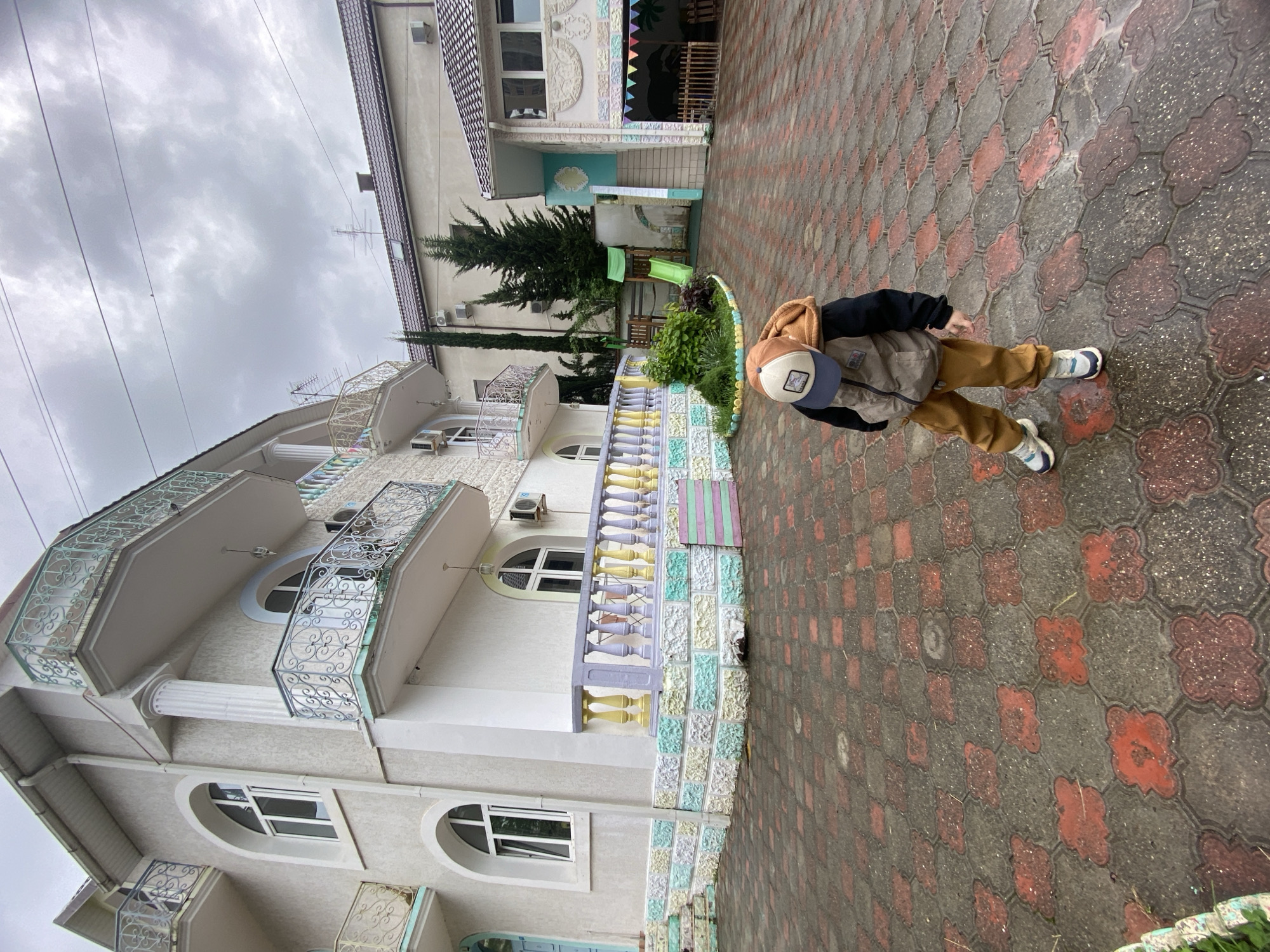 The image shows a child standing on a paved walkway beside a building with balconies. The building is a light color with decorative details, including arched windows and colorful trim. A second building is visible in the background. The sky is overcast.
Based on the architectural style and the color scheme of the buildings, it is likely that the location is in the city of Sochi, Russia.