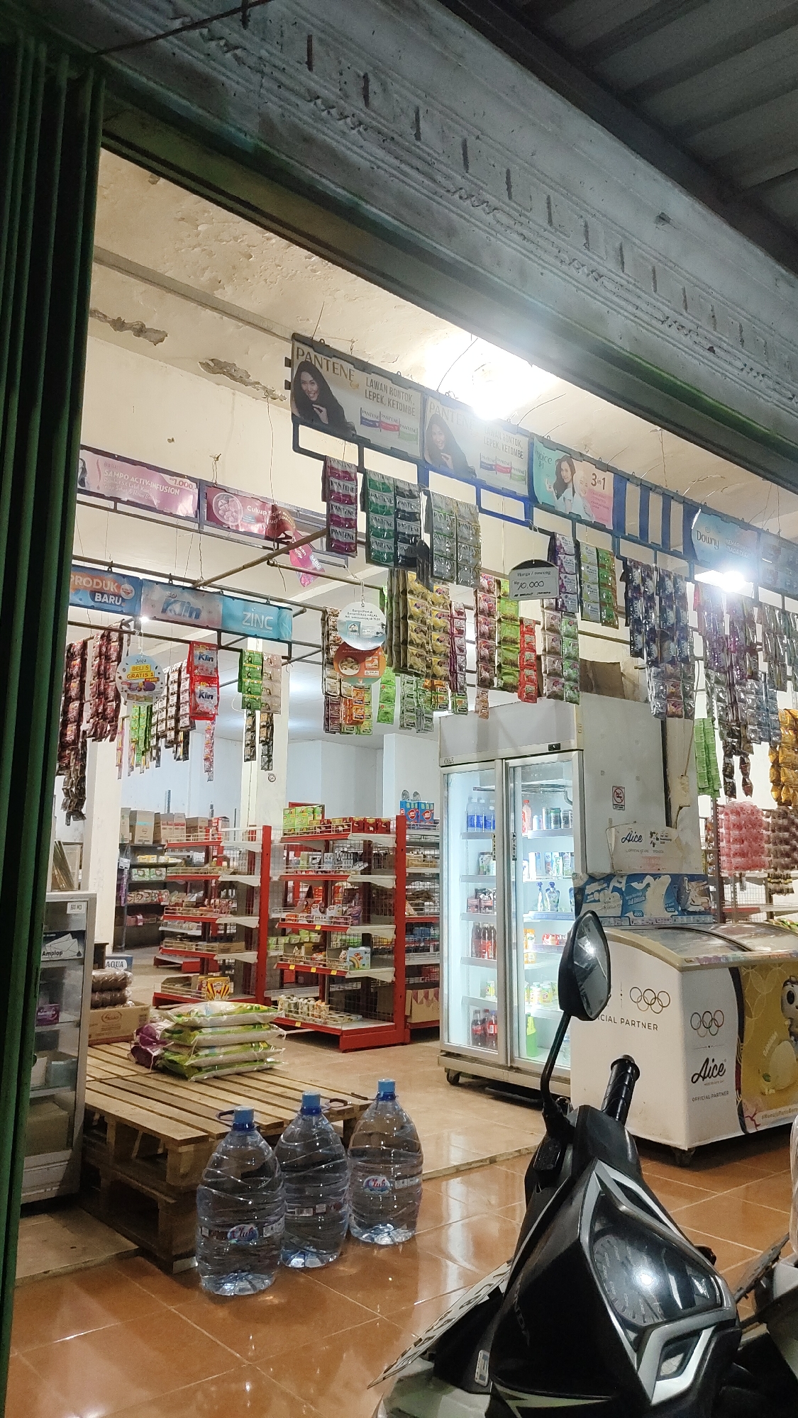 This image captures the interior of a small convenience store, likely in Malang, Indonesia, judging by the script and context. The store appears to be well-stocked with a variety of packaged goods, including what seem to be food items, toiletries, and beverages, arranged on shelves and hanging displays. Prominently featured are large jugs of drinking water near the entrance, and several display refrigerators. A motorcycle is parked in the foreground, partially obscuring the view of the store's interior. The lighting suggests it might be evening or a dimly lit interior. The overall impression is of a typical local shop, bustling with merchandise ready for sale.