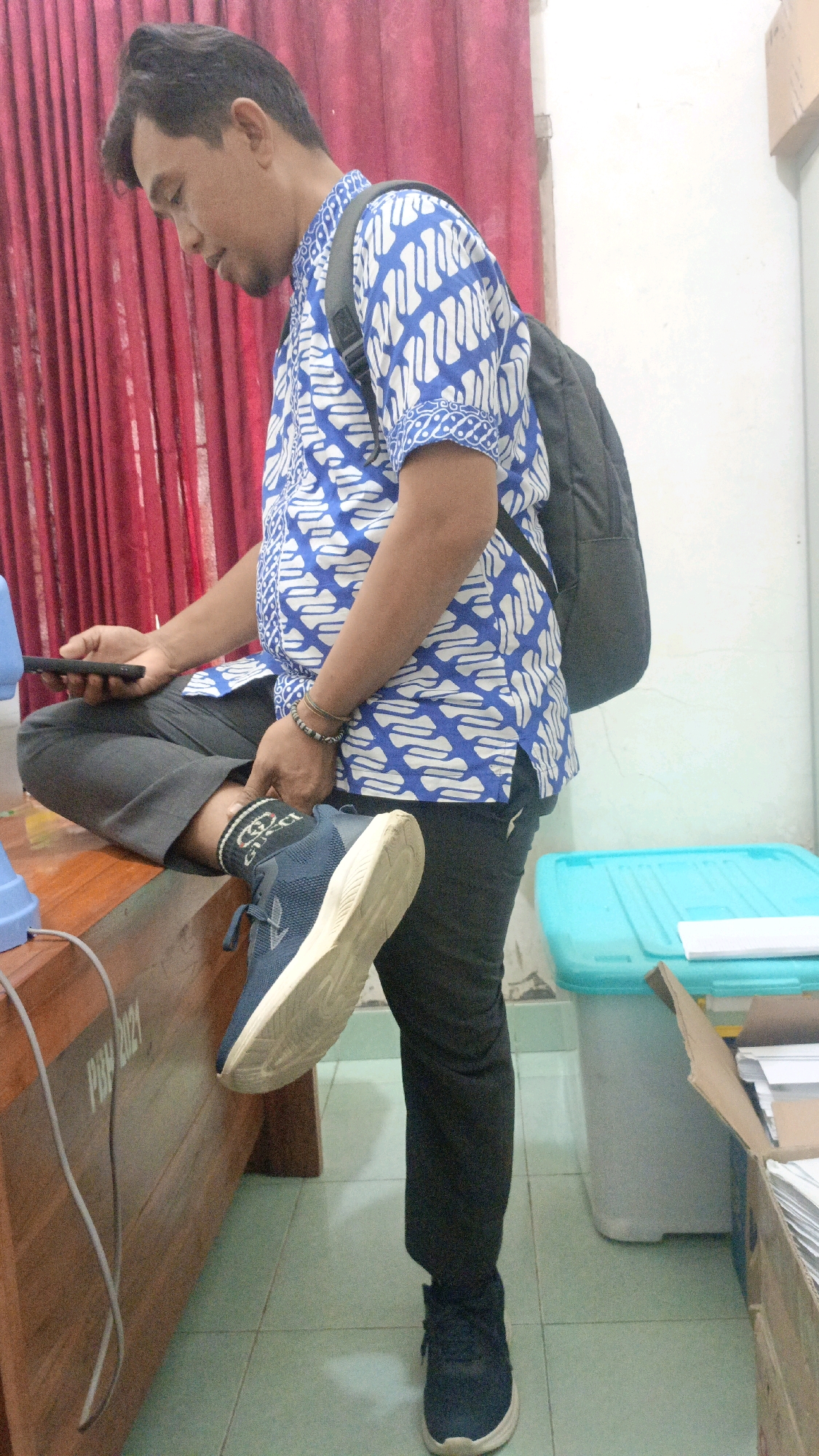A man is seated on a wooden desk, holding a dark smartphone in his left hand and a black backpack is slung over his right shoulder. He is wearing a blue and white batik print shirt, dark gray trousers, and dark blue athletic shoes with white soles. His left leg is crossed over his right, with his left foot resting on the edge of the desk. His right leg is extended downwards, with his right foot on the floor. The man appears to be looking down at his phone.
The background shows a red curtain, a light blue plastic bin, and a cardboard box filled with papers. The floor is tiled in a light green color. The environment suggests an indoor setting, possibly an office or a classroom. The text "PBM 2021" is visible on the side of the wooden desk, indicating a potential date or affiliation.
The image was taken in Taliwang, Indonesia, likely in a room with basic furnishings. The lighting is moderate, with a slight shadow cast on the floor by the man. The overall scene is casual, with the man appearing relaxed as he uses his phone.