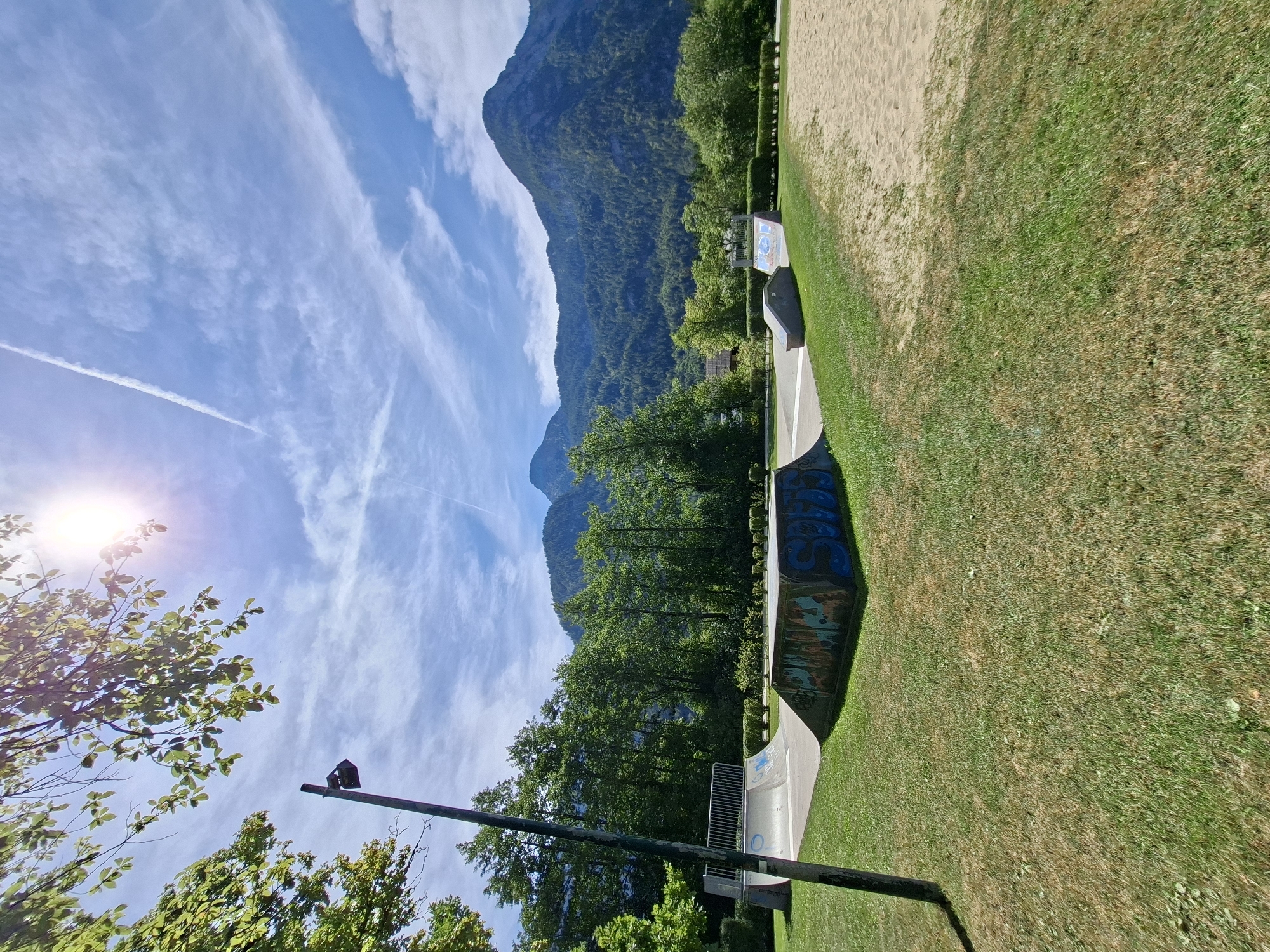 The image depicts a skatepark in Obertraun, Austria, bathed in bright sunlight under a partly cloudy blue sky. In the foreground, a green grassy area transitions into a sandpit on the right. The skatepark features several concrete ramps, one of which is covered in colorful graffiti. A tall, dark lamppost stands to the left of the park. Behind the skatepark, a dense line of lush green trees forms a backdrop, beyond which majestic, tree-covered mountains rise, their peaks partially obscured by the sky. A contrail from an aircraft is visible streaking across the sky. The sun, positioned in the upper left quadrant, casts a lens flare onto the image.