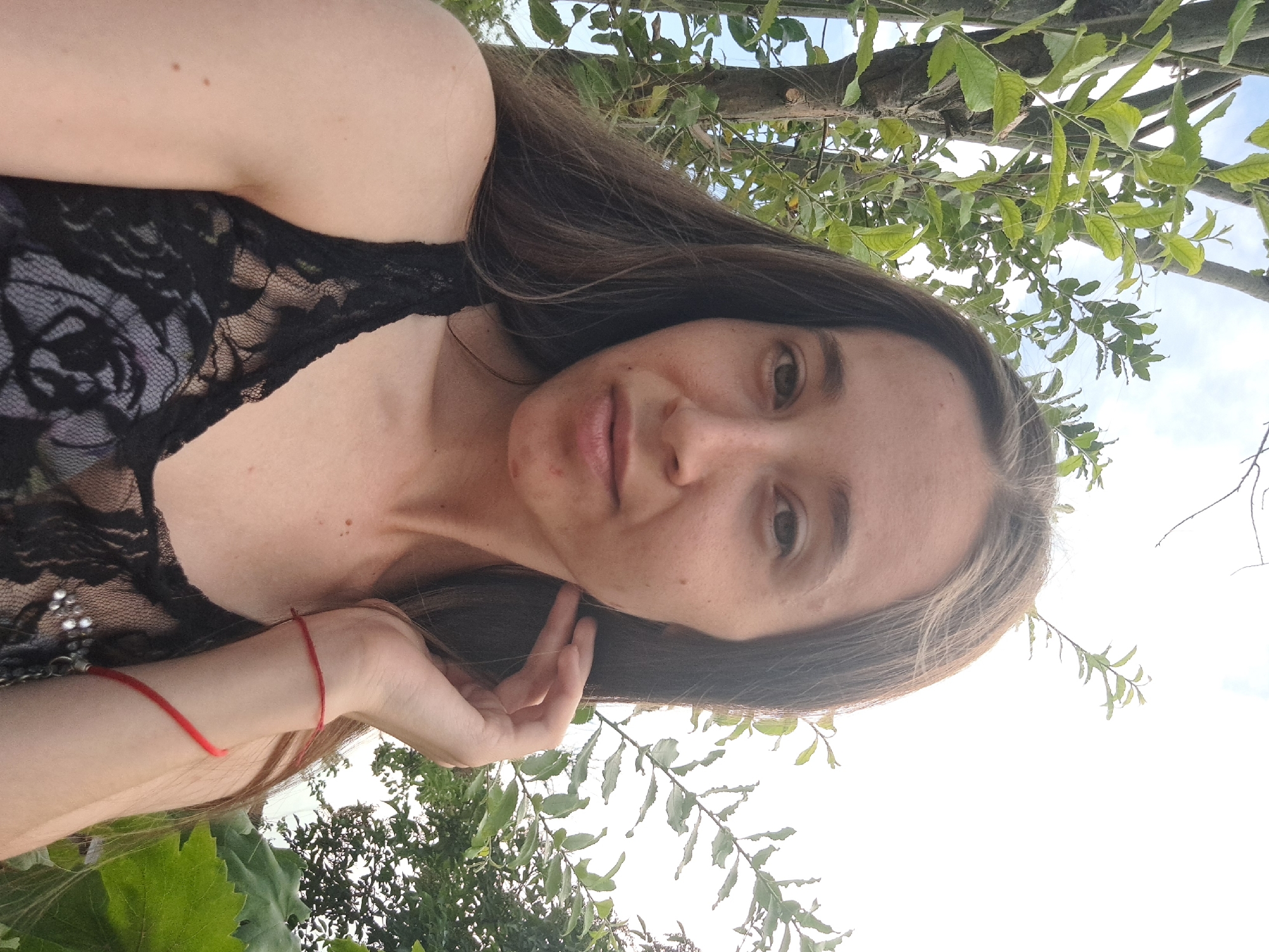 A young woman with long, brown hair and fair skin is captured in a selfie outdoors. She is wearing a black lace top. Her right hand is raised to her face, touching her jawline. A thin red string bracelet is visible on her left wrist. The background consists of green foliage and a bright sky, suggesting a natural setting, possibly a garden or park. The image does not provide information about the specific location within Vypasne, Ukraine.