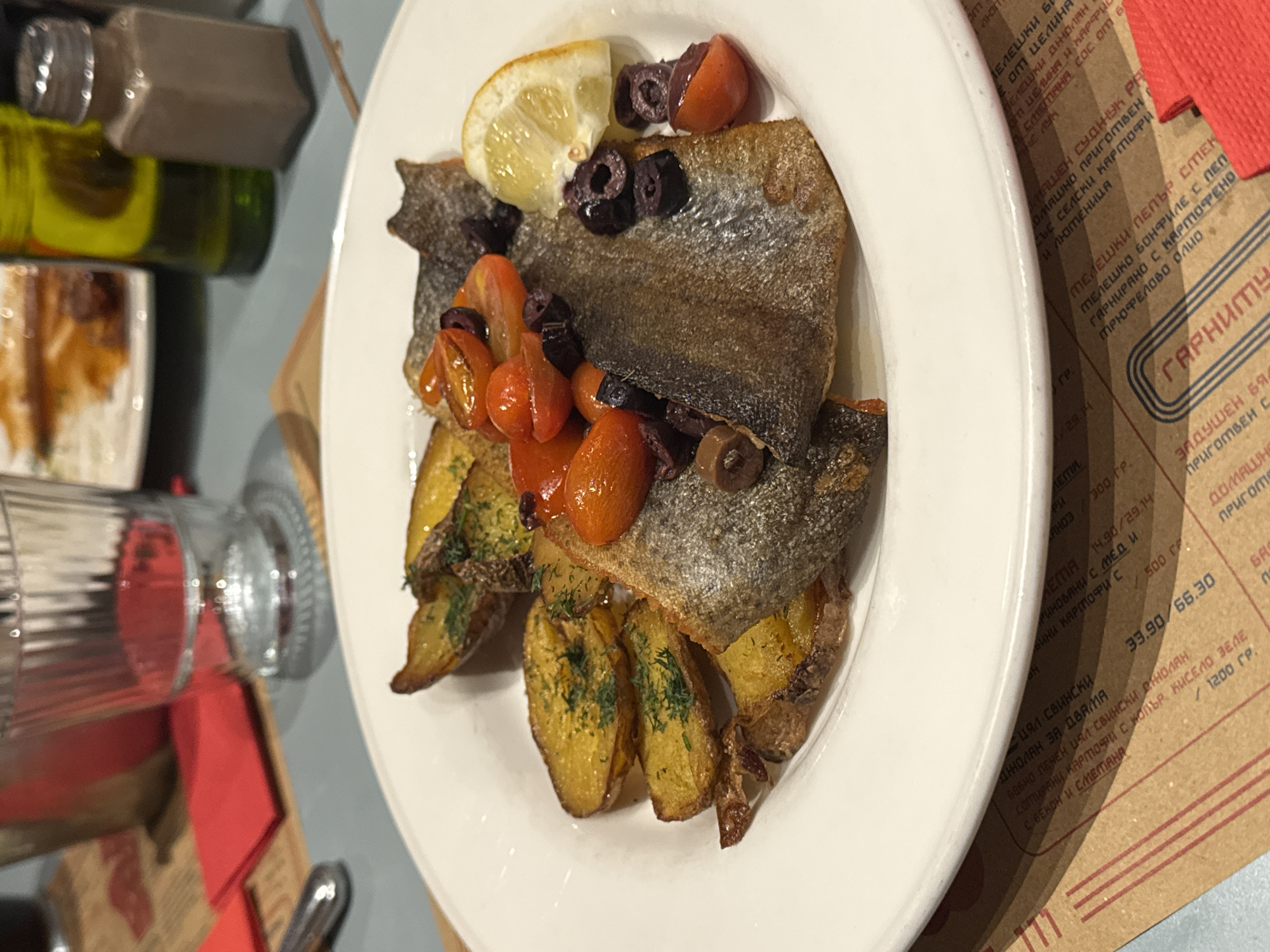 The image shows a plate of food, likely taken indoors, on a table in a restaurant, potentially in Sofia, Bulgaria, based on the Cyrillic script on a menu/placemat near the plate. The main dish appears to be pan-fried fish with skin on, served over roasted potatoes seasoned with dill and topped with blistered cherry tomatoes, sliced black olives and a slice of lemon. Other visible objects include glass bottles, tableware, a menu or placemat, and a glass.