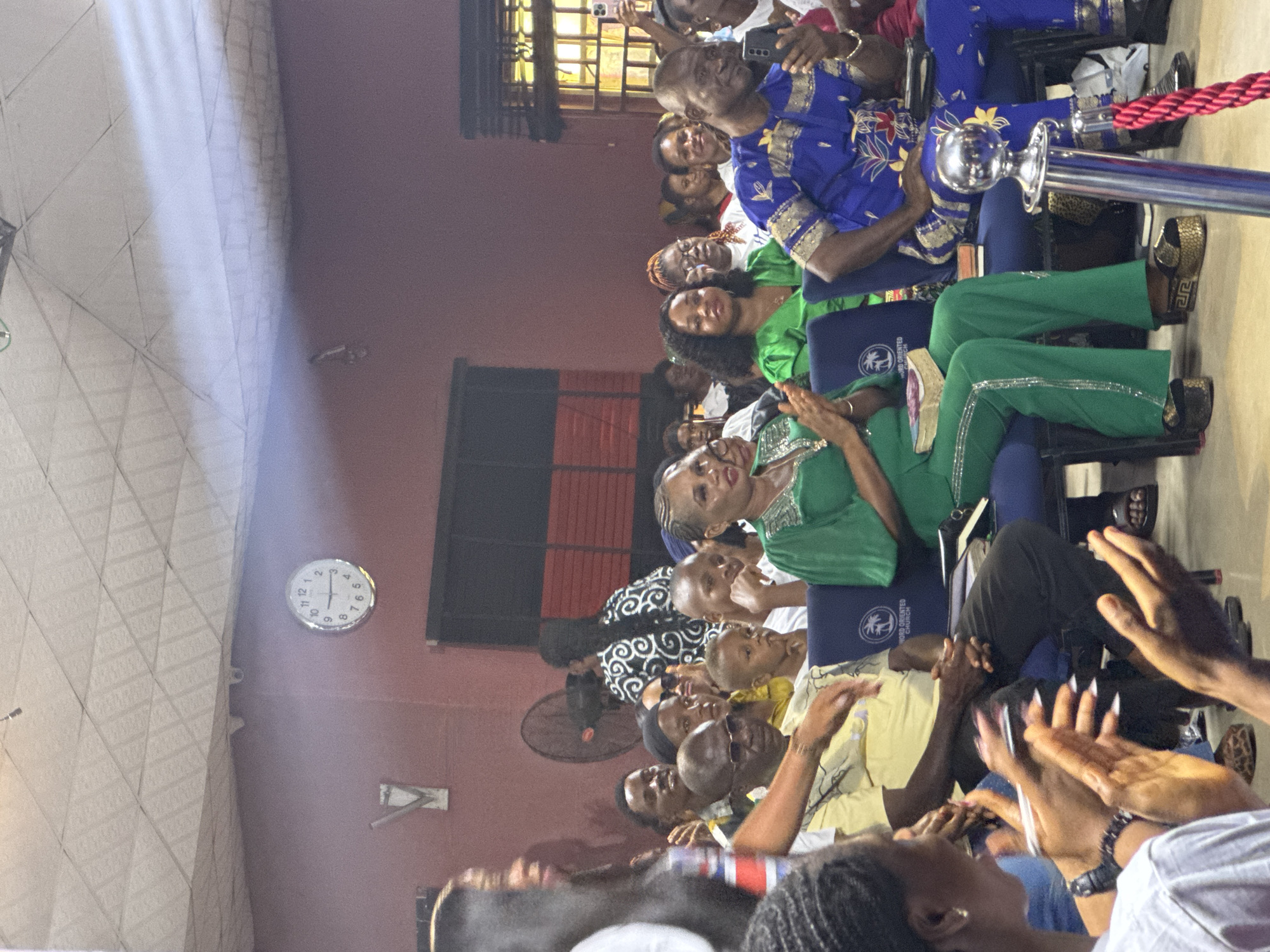 Here is a forensic description of the image.
The image shows an interior scene, likely in Nkpor, Nigeria, featuring rows of people seated and engaged in a gathering. The individuals are predominantly African, of various ages, and are dressed in colorful attire, including green and blue garments. Many are clapping, suggesting an atmosphere of appreciation or celebration.
A silver stanchion with a red rope is visible near the front row. Several individuals are holding smartphones, potentially recording or photographing the event. A wall-mounted clock indicates the time. There's also a wall-mounted fan in the background. The "Word Oriented Church" logo is visible on a blue document.