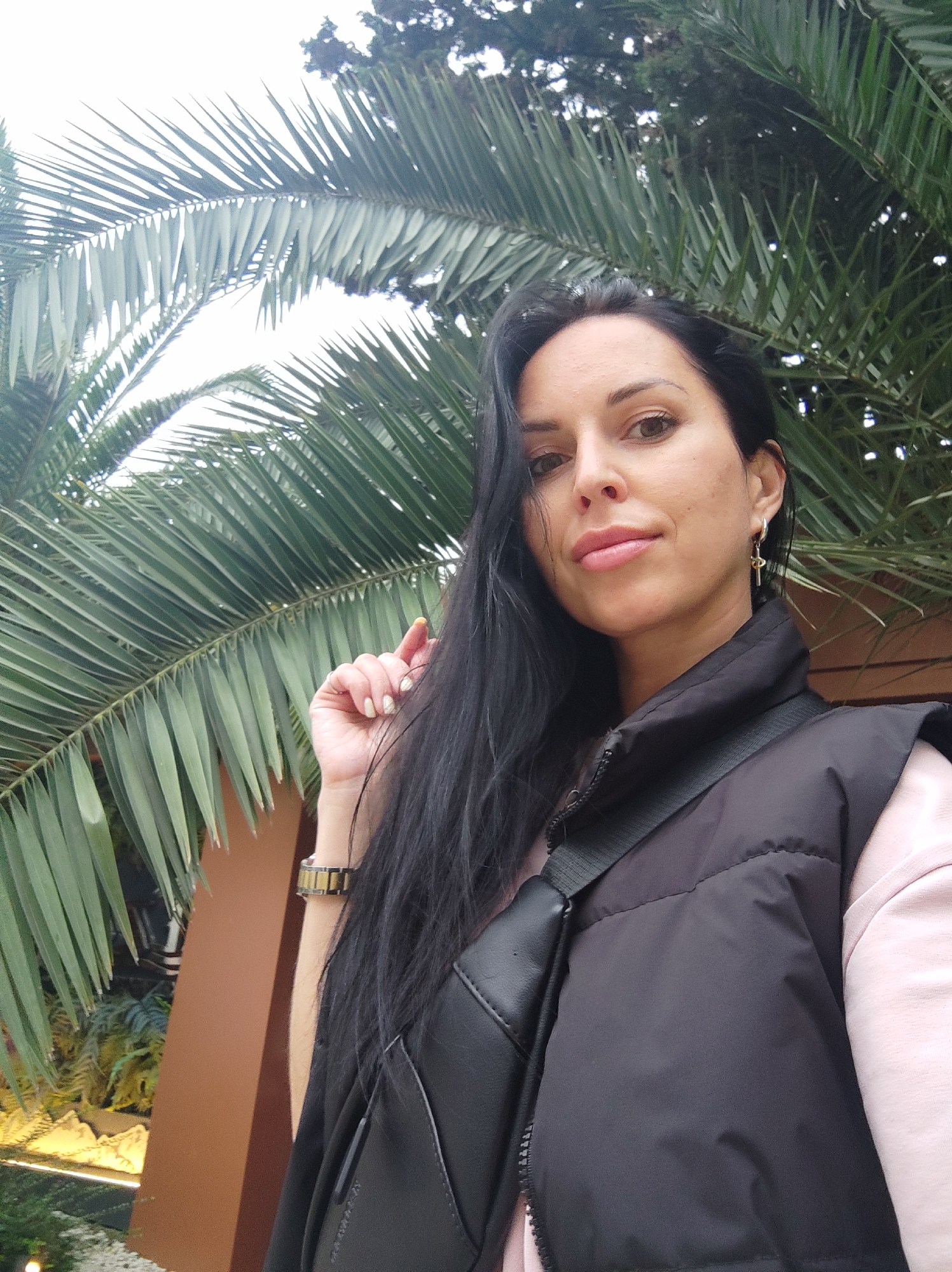 A medium shot captures a woman with long, black hair against a backdrop of lush, green palm fronds. She wears a black vest over a light pink garment, with a black shoulder bag slung across her chest. A gold wristwatch adorns her left wrist, and dangling earrings are visible. The location is Sochi, Russia, indicated by the presence of palm trees and architectural elements. The woman is holding her hand up near her face. The overall ambiance suggests an outdoor setting, possibly a park or garden.