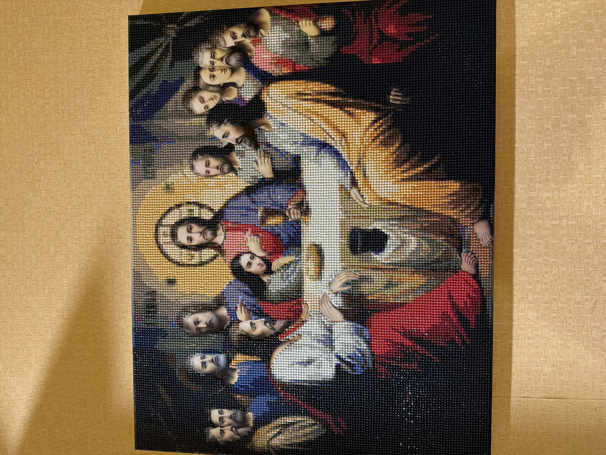 The image shows a diamond painting of Leonardo da Vinci's "The Last Supper." The painting depicts Jesus Christ and his twelve apostles gathered around a table for their final meal. The diamond painting is framed and hung on a textured, light brown wall. Shadows are cast on the wall from the top left, suggesting an overhead light source. The overall scene is an indoor still life. There is no information in the image to suggest its location in Kyiv, Ukraine.