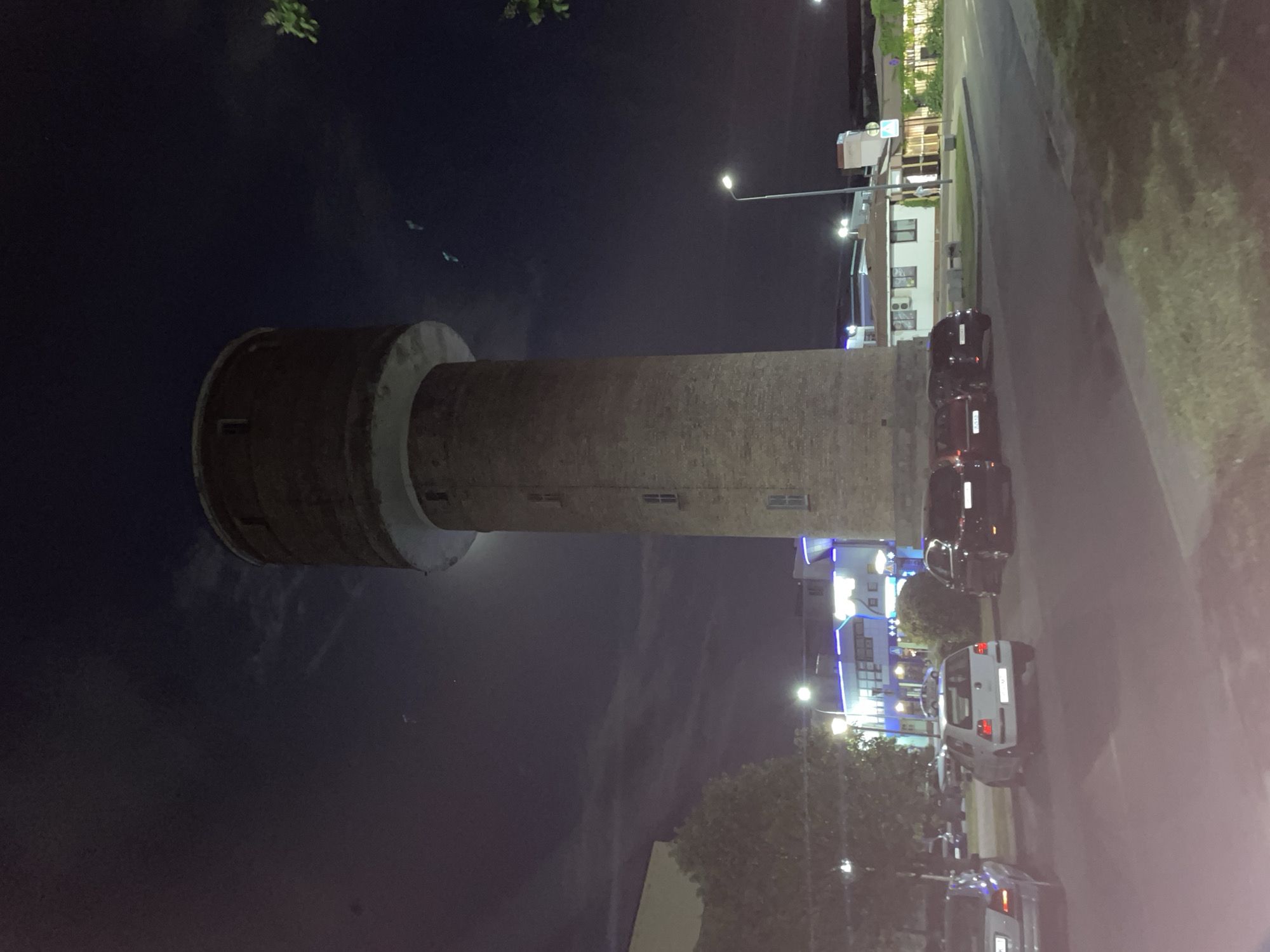 Here's a detailed description of the image:
The image shows a brick water tower at night in Brest, Belarus. It is a tall cylindrical structure with a wider section at the top. There are small windows visible along the tower's shaft.
To the right of the tower is a street with several parked cars. There are buildings and streetlights lining the road. The sky is dark, and the overall lighting suggests it is nighttime. Vegetation is present on the side of the road. There are no people visible.