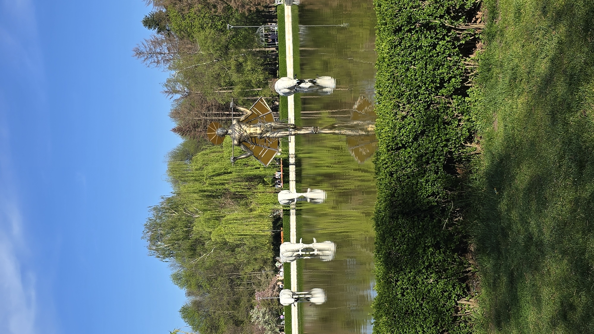 This image captures a vibrant and serene park landscape, likely in Sofia, Bulgaria, featuring a long, reflective pond bordered by lush greenery and striking sculptures under a clear blue sky.
The central focus is a large, golden-toned contemporary statue positioned on the left bank of the pond. This impressive sculpture depicts a human-like figure, possibly male, adorned with large, geometric, wing-like structures extending from its back, and a halo-like disc around its head. The figure holds what appears to be a staff or spear. Its detailed form and reflection are prominently visible in the calm water.
Lining the pond at regular intervals are several smaller, abstract white sculptures. These modern art pieces are partially submerged or situated directly in the water, creating a rhythmic visual sequence and casting their own distinct reflections.
On the left side of the pond, a white paved pathway runs alongside, flanked by a variety of trees. These include tall deciduous trees with light green, fresh foliage, and some darker evergreens. Willow-like trees with their characteristic drooping branches also add to the verdant setting. In the distance, a few indistinct figures of people can be seen enjoying a stroll.
The right side of the pond is bordered by a meticulously trimmed, dense green hedge, beyond which lies a wide expanse of manicured green grass. Shadows are cast across the grass, indicating bright, direct sunlight. The sky overhead is a vibrant, clear blue with minimal cloud cover, suggesting a pleasant, sunny day, likely late morning or early afternoon. The scene conveys a sense of tranquility and artistic beauty within a well-maintained natural environment. There is no visible text in the image.